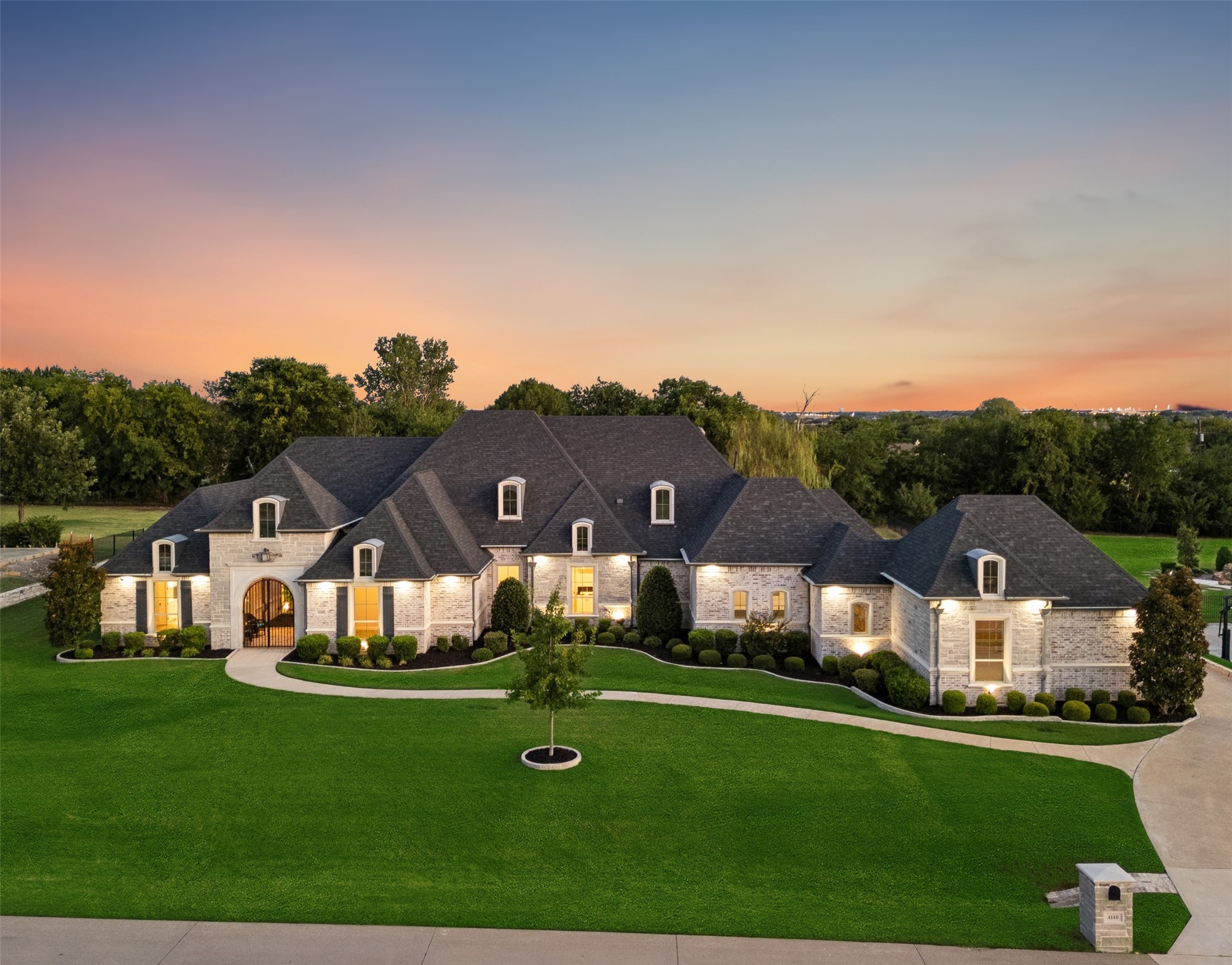 Large stone home with illuminated windows, expansive lawn, and curved driveway at dusk.
