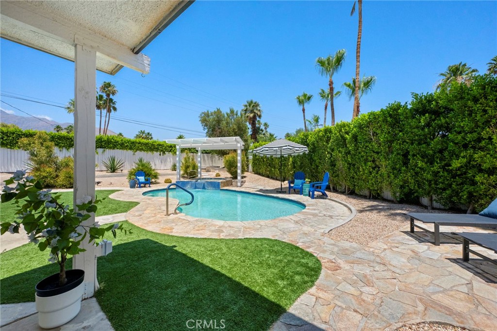 Backyard swimming pool bordered by a stone patio, green lawn, white pergola, and tall hedges.