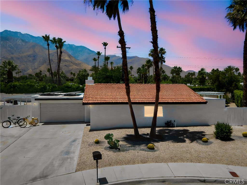 Single-story white home with terracotta tile roof, gravel yard, tall palms, and mountains at dusk.