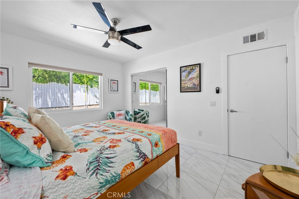 Furnished bedroom with a colorful floral quilt, ceiling fan, and large windows overlooking a fenced yard.