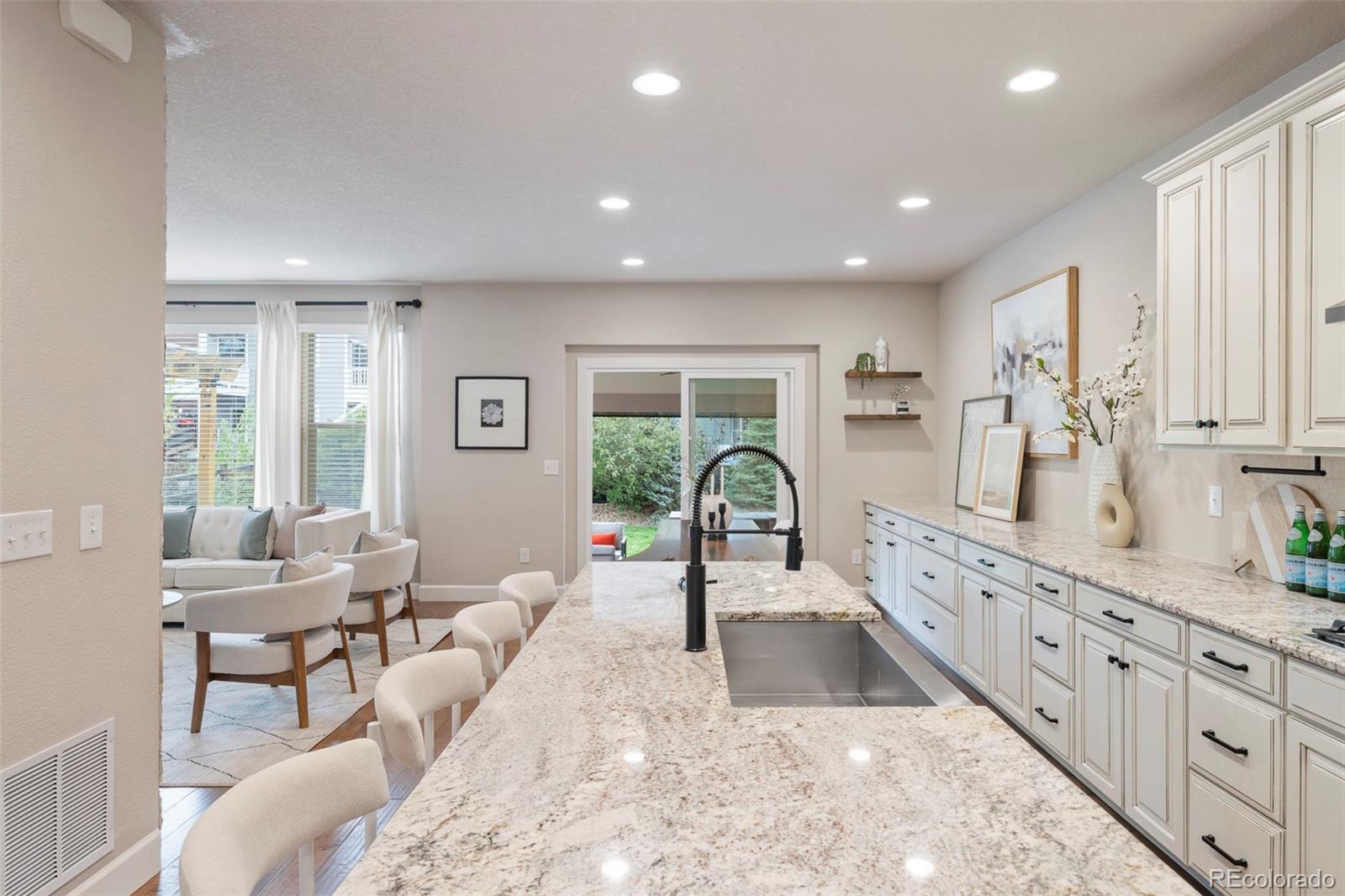Bright kitchen with a long stone island and black faucet, opening to a sitting area and garden.