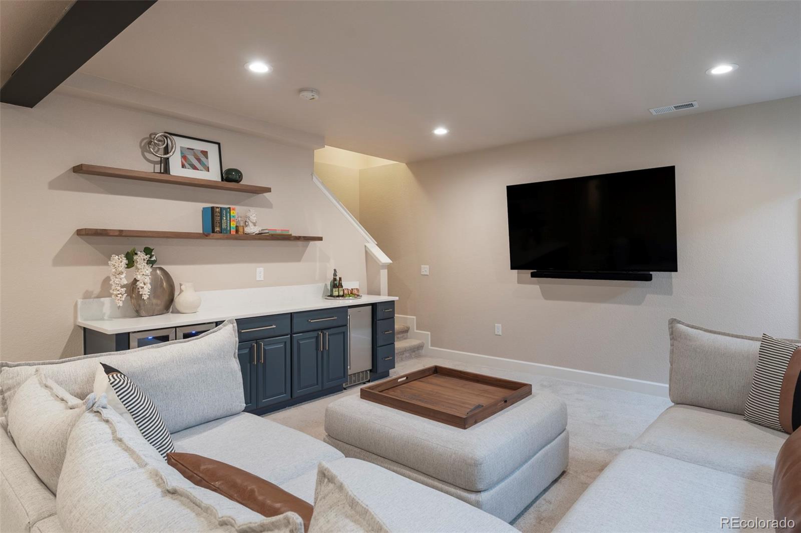 Lower-level living room with a wall-mounted TV, blue-cabinet wet bar, floating shelves, and sectional seating.