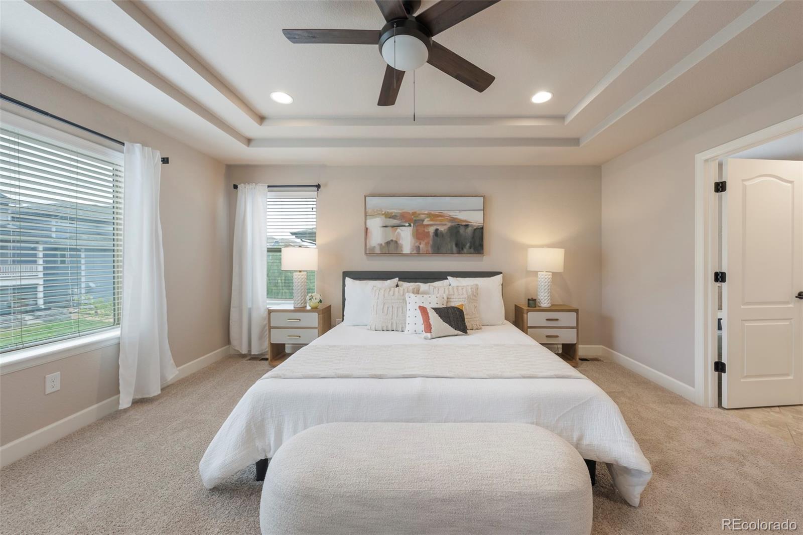 Staged bedroom with a tray ceiling, dark ceiling fan, and large windows beside the bed.