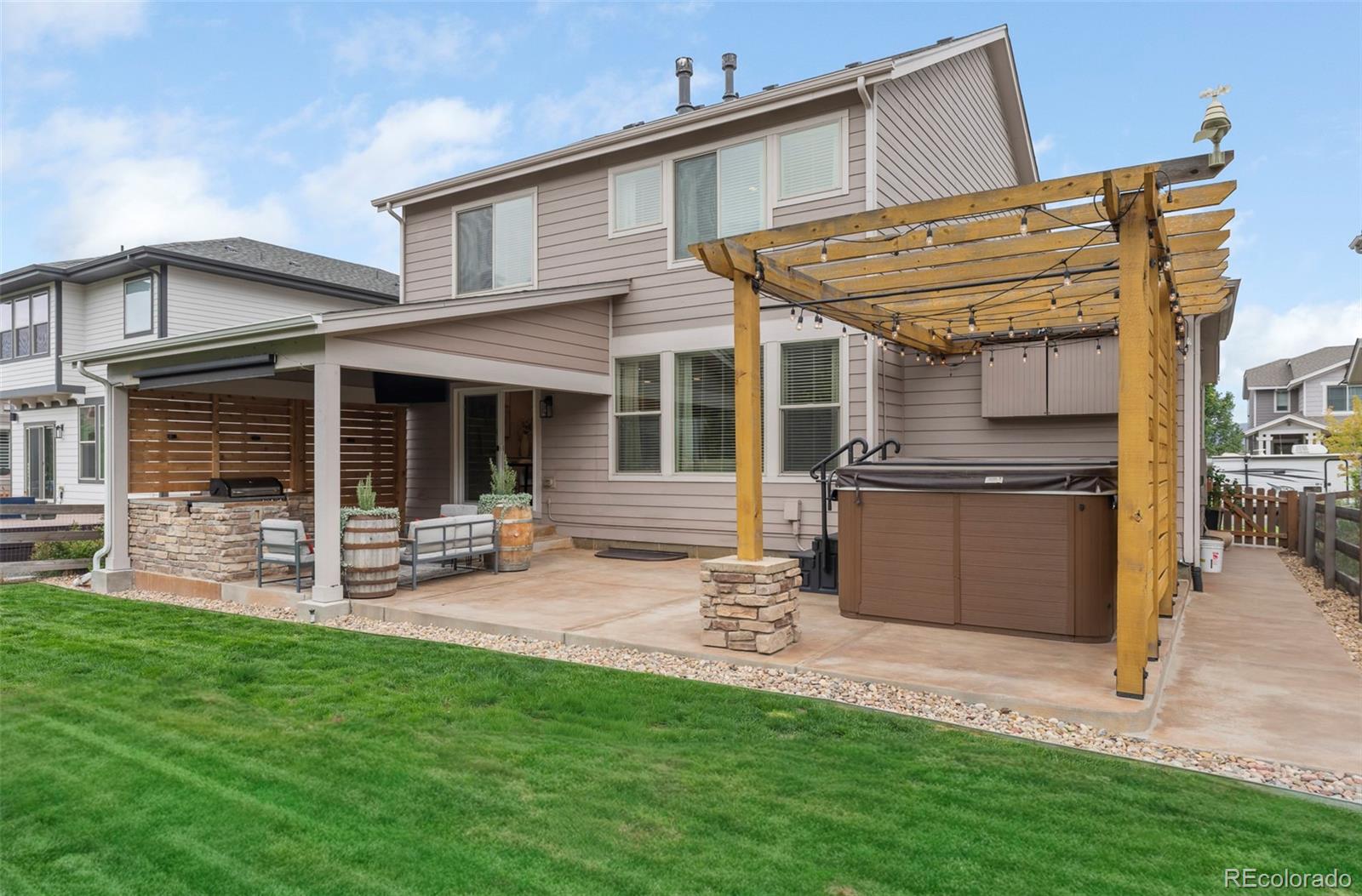 Home’s backyard patio with a wooden pergola, hot tub, stone outdoor kitchen, and green lawn.