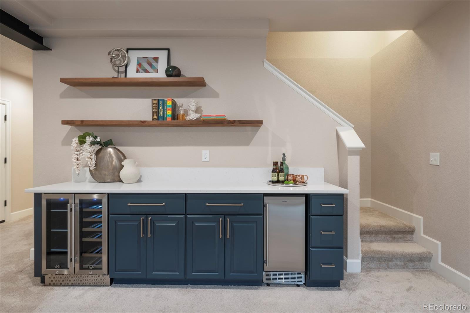 Basement wet bar with navy cabinets, two beverage refrigerators, floating wood shelves, and nearby stairs.