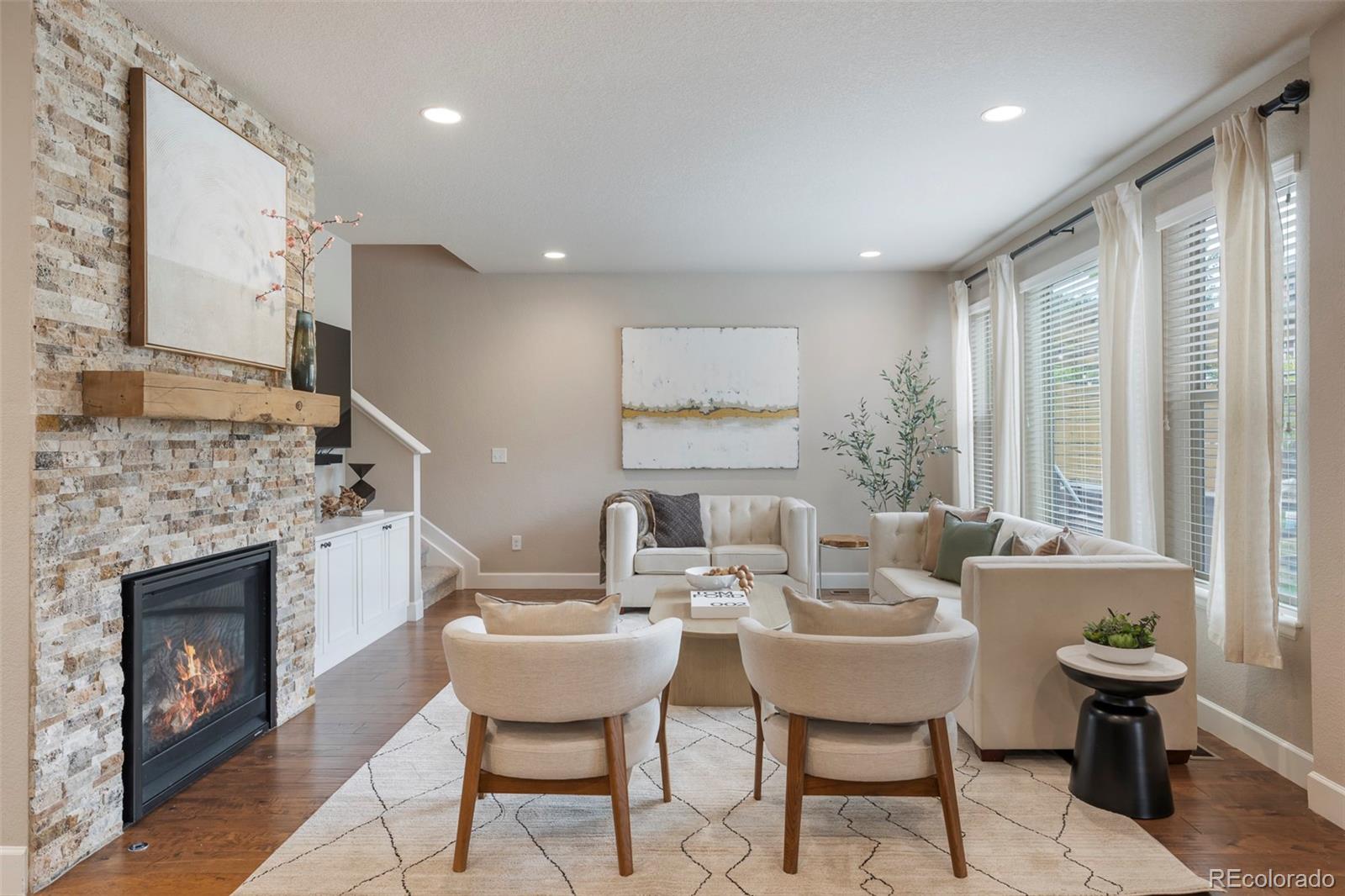 Staged living room with a stone fireplace and neutral seating beside large windows.