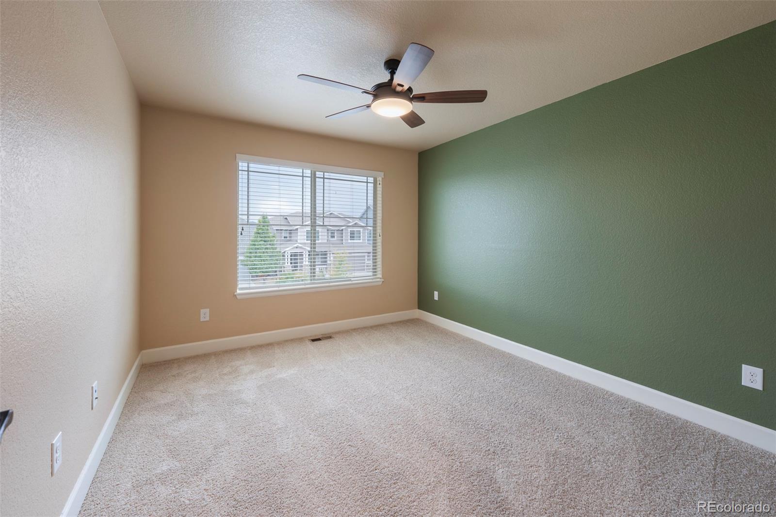 Empty carpeted bedroom with a green accent wall, ceiling fan, and window overlooking neighboring homes.