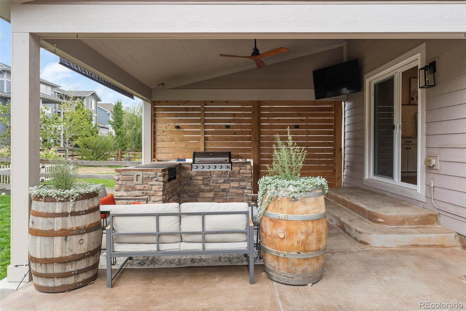 Covered patio with stone grill island, wood privacy screens, ceiling fan, and barrel planters.