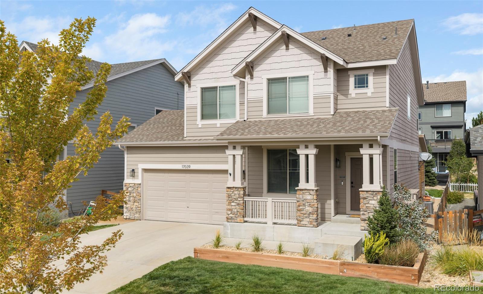 Two-story beige house with stone porch columns, a two-car garage, and raised front garden beds.