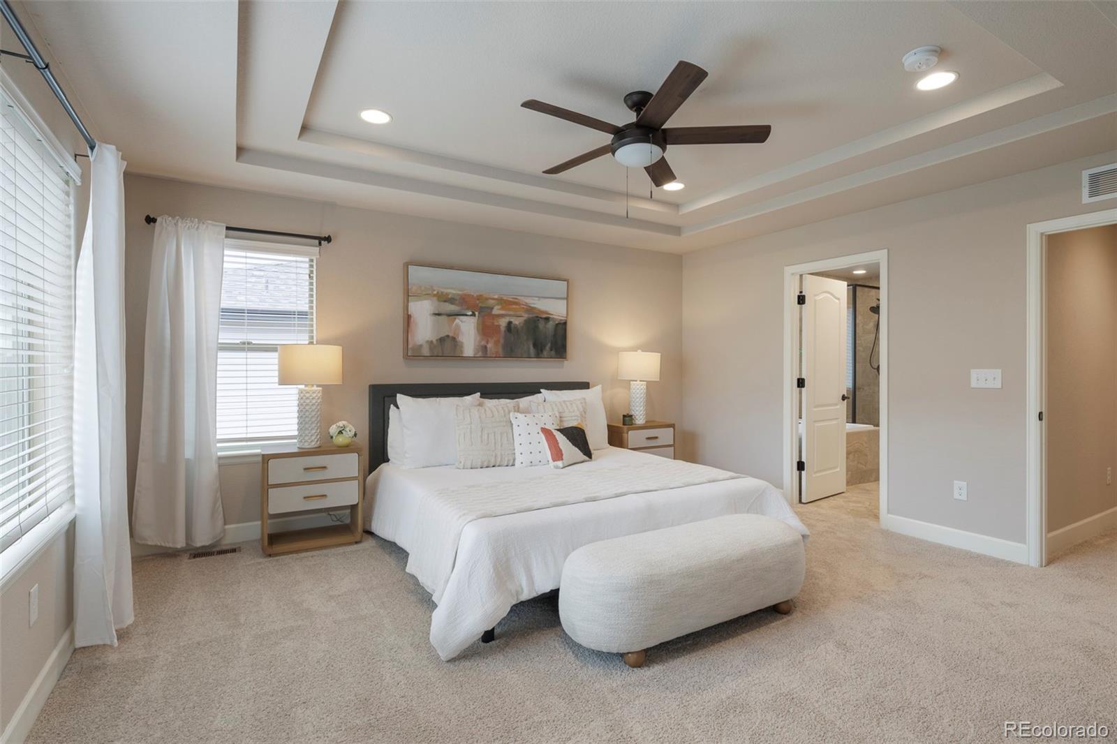 Staged bedroom with a king bed, upholstered bench, tray ceiling, and open doorway to the ensuite bathroom.