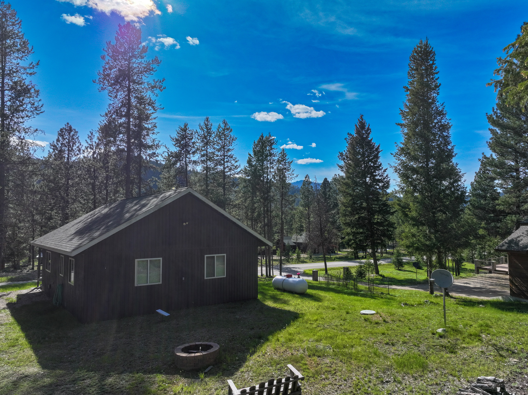 Dark-brown cabin in a conifer-lined yard with a circular fire pit and propane tank.