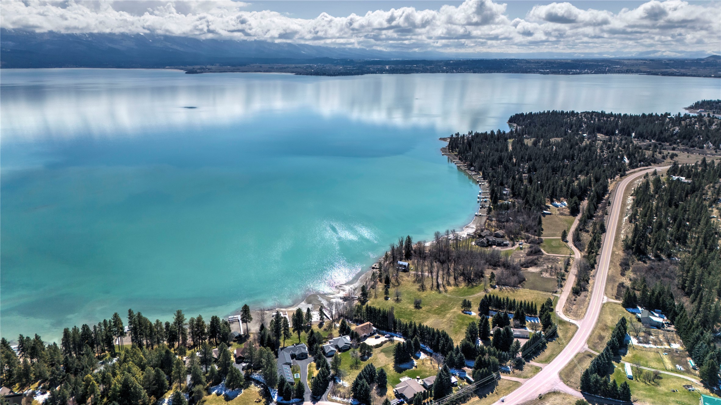 Aerial lakeside neighborhood beside turquoise water, with wooded shorelines and snow-dusted mountains.