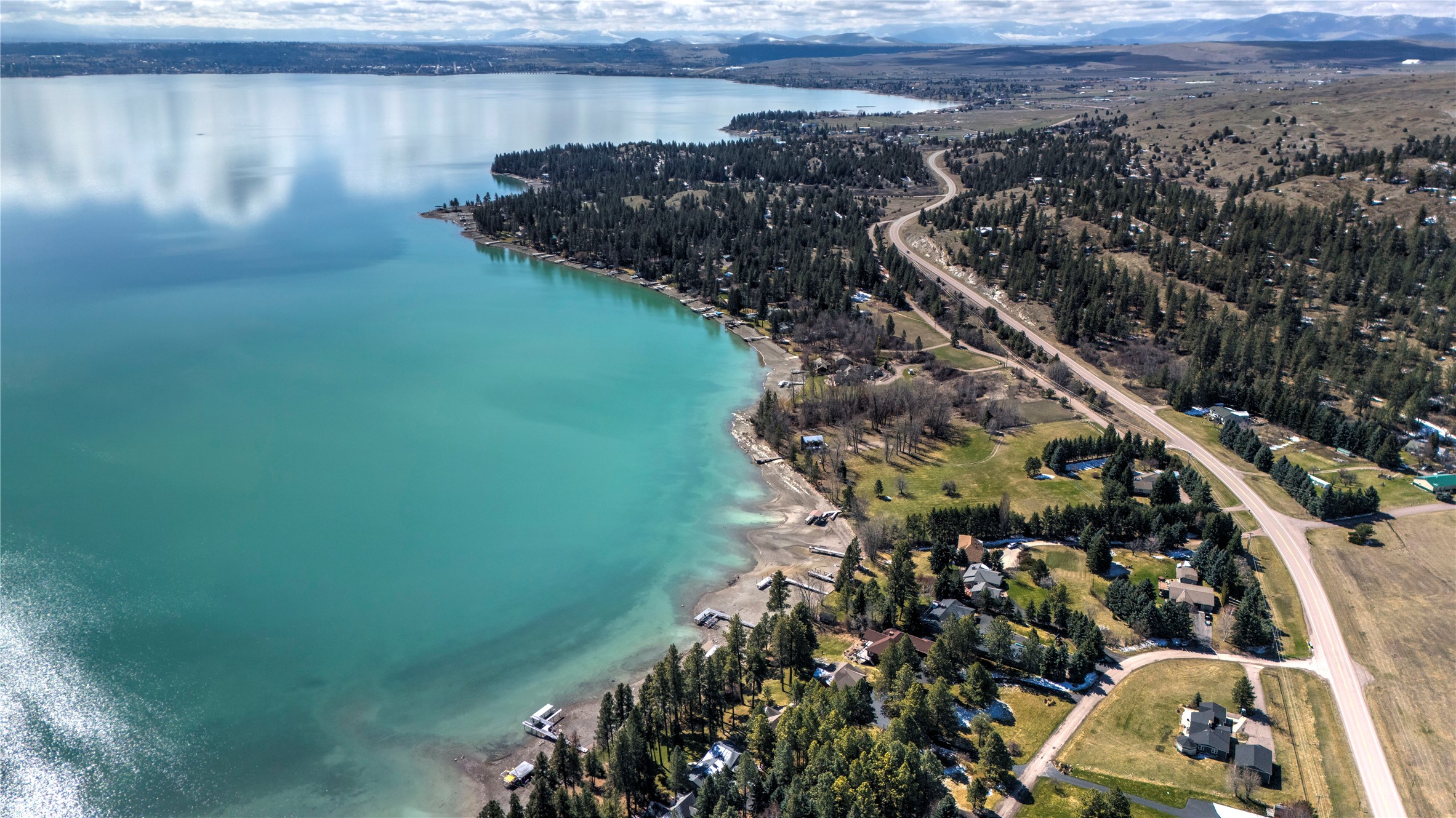 Aerial view of lakeside homes and winding road beside turquoise Flathead Lake, with mountains in the distance.
