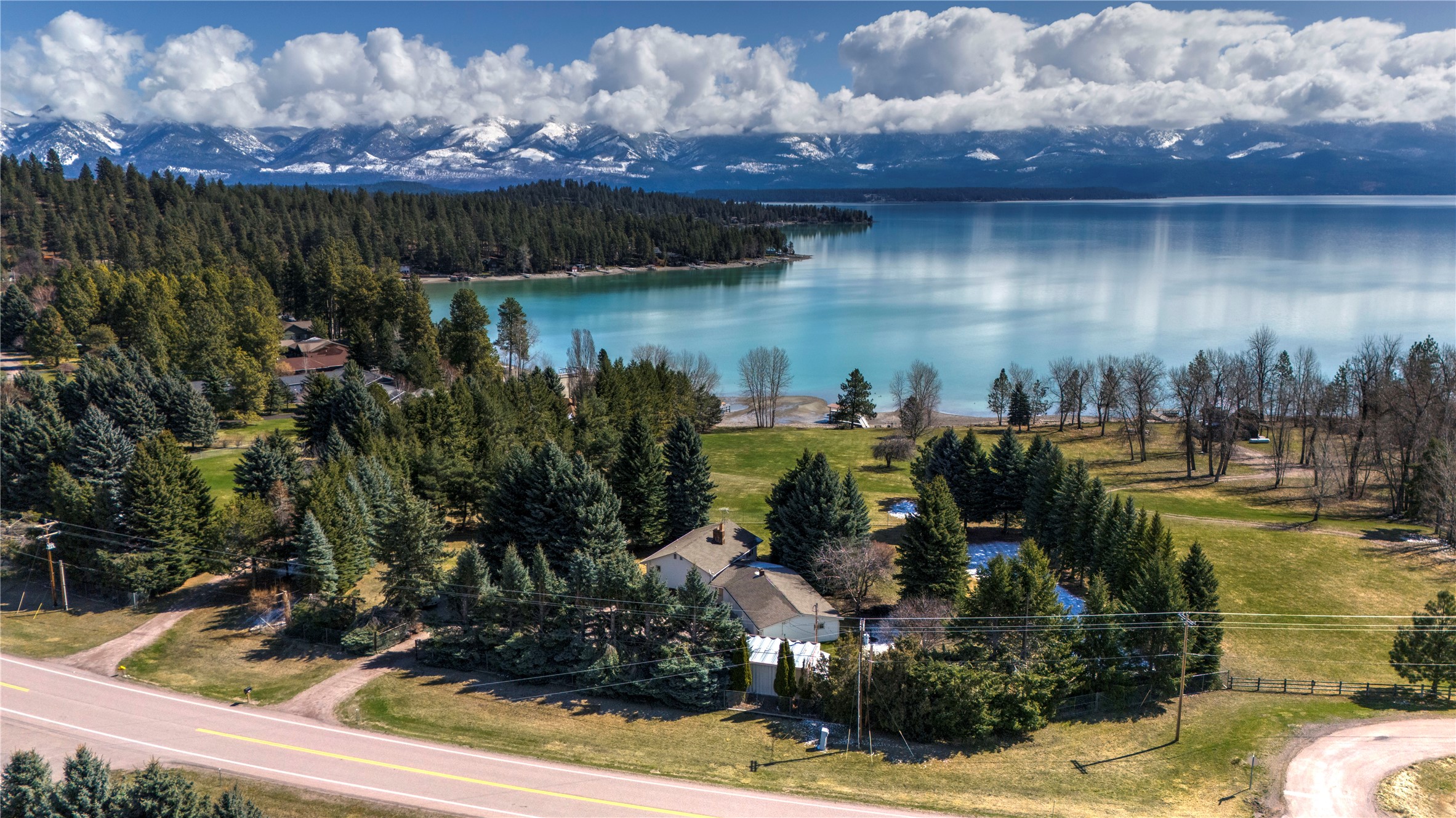 Aerial view of a tree-lined lakeside home with calm water and snow-dusted mountains beyond.