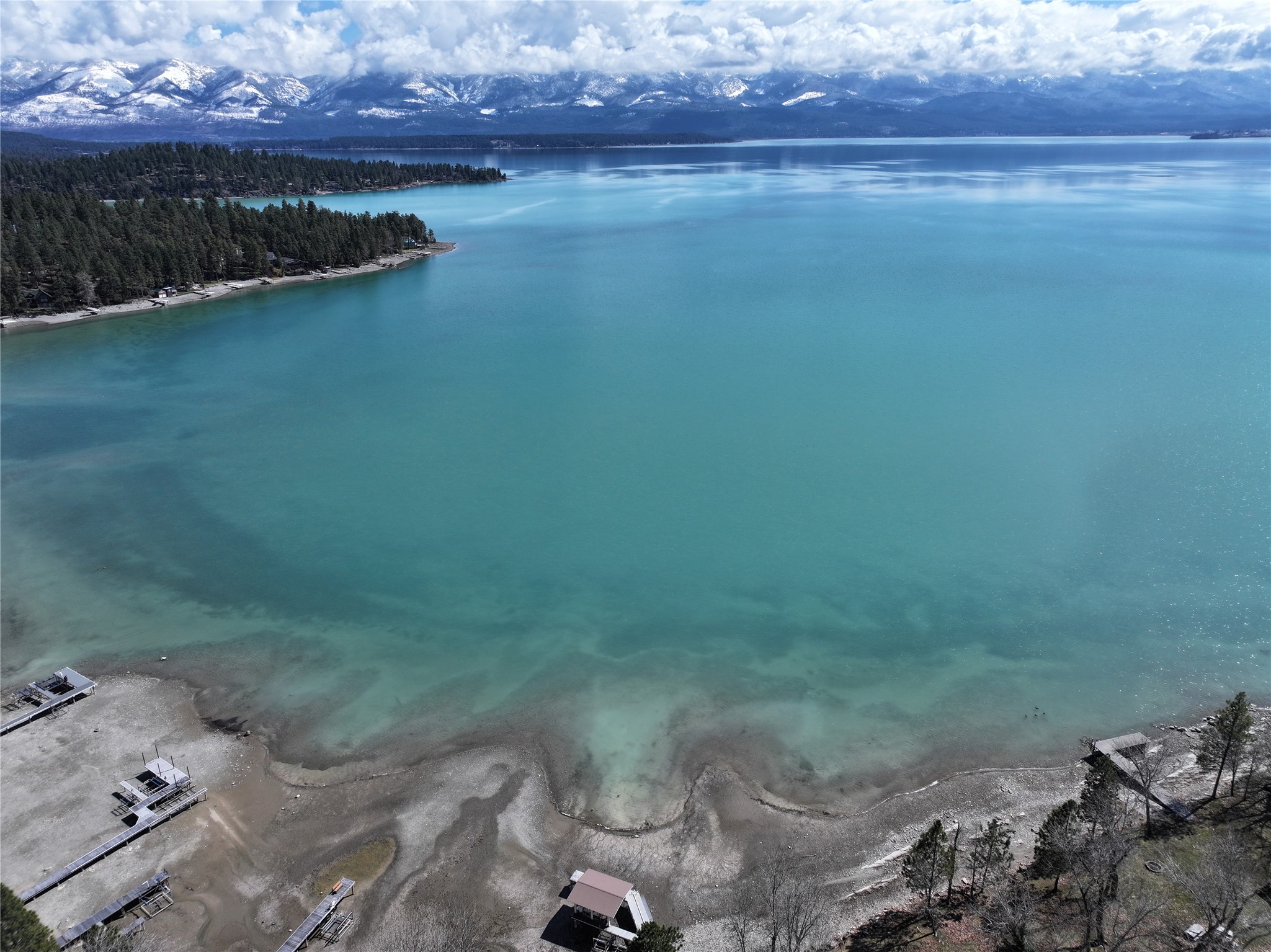 Aerial view of a turquoise lake, rocky shoreline with docks, wooded peninsula, and snow-covered mountains.