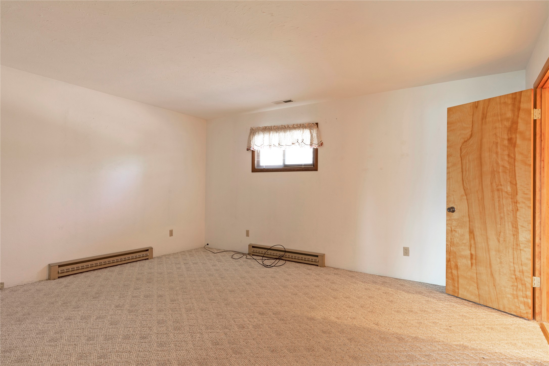 Empty carpeted room with baseboard heaters, a small curtained window, and an open wooden door.