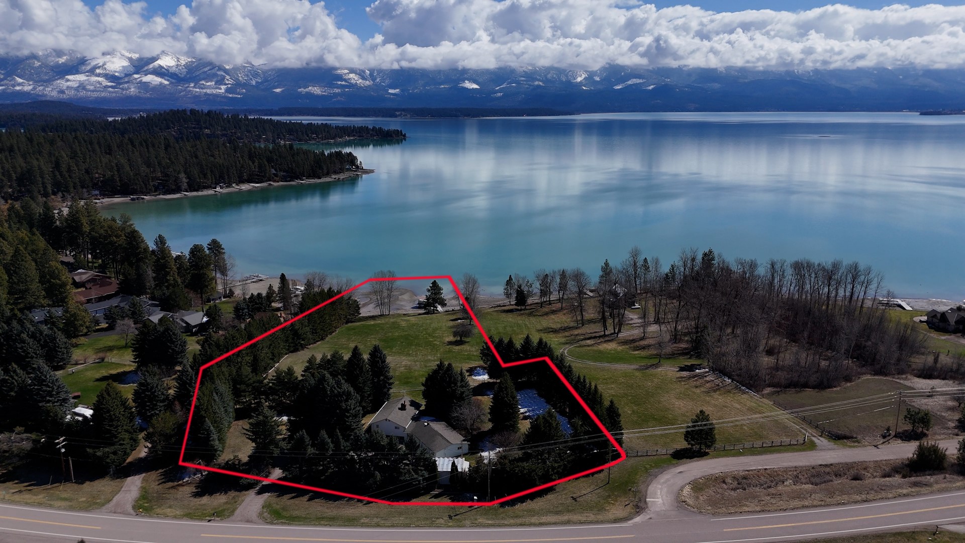 Aerial view of a lakeside home and lawn outlined in red, with snow-dusted mountains across the water.