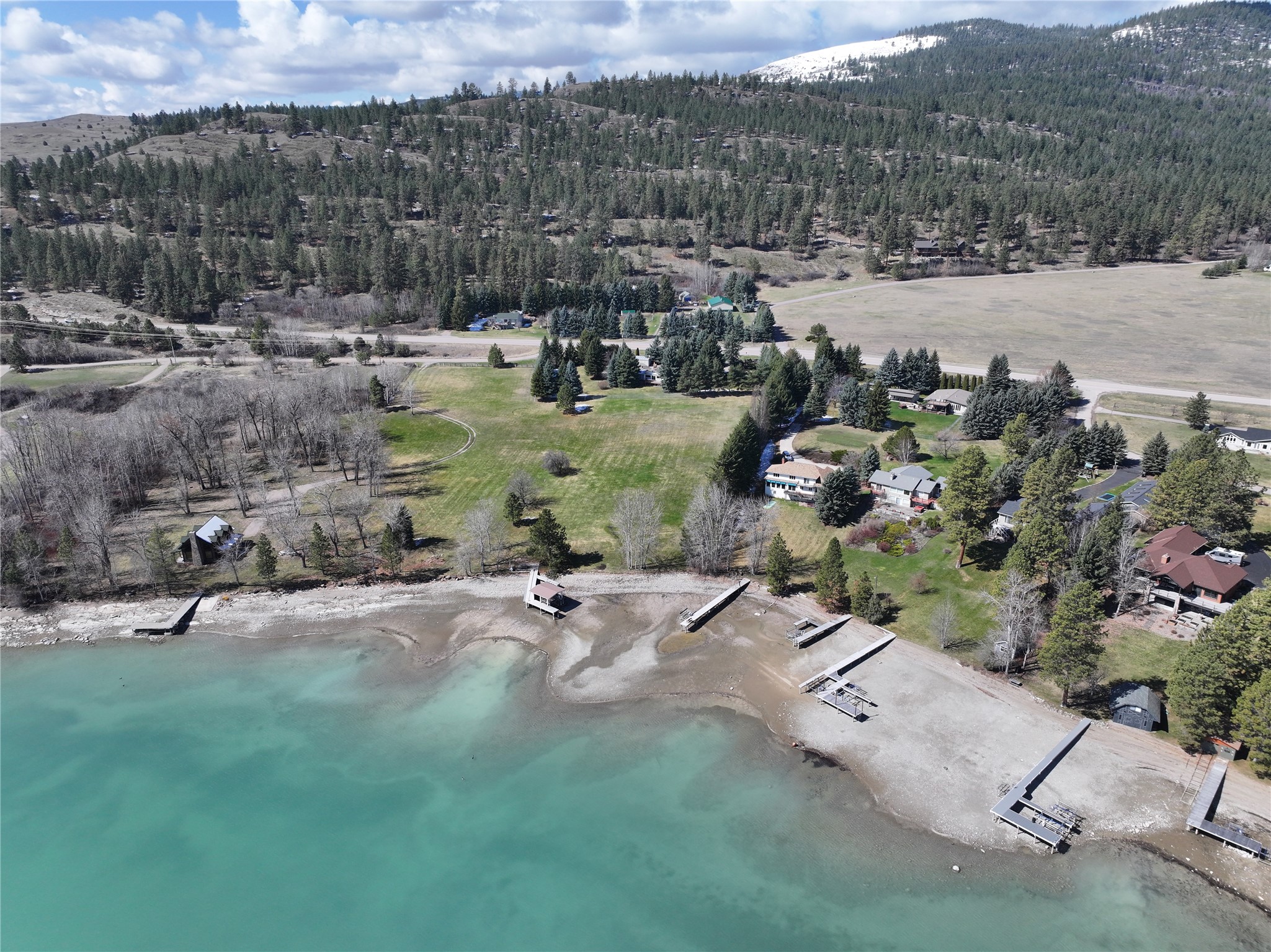 Aerial view of lakeside homes and several docks along turquoise water, backed by a wooded hillside.