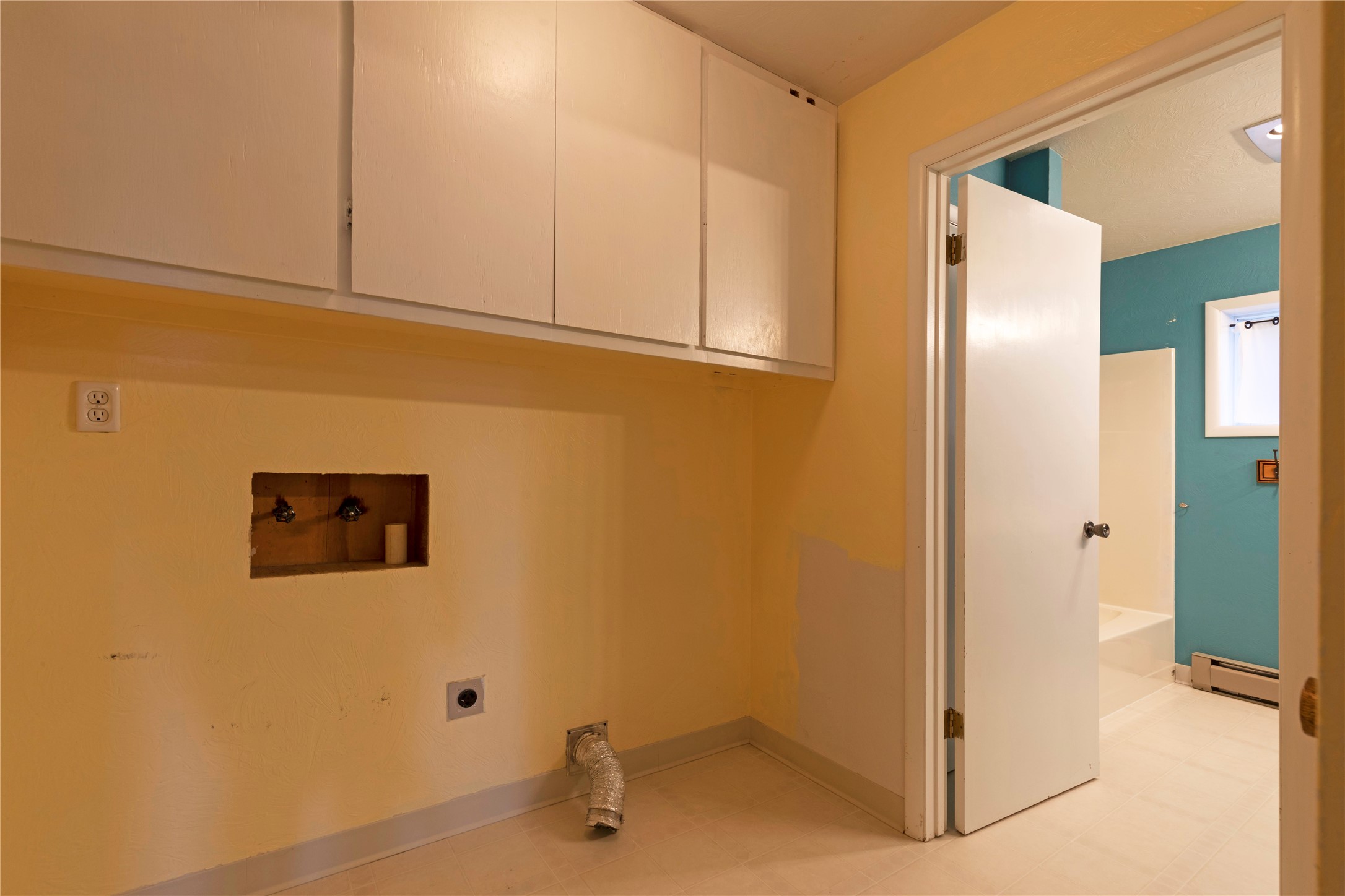 Empty laundry area with overhead cabinets, exposed hookups, and a dryer vent hose beside a teal bathroom.