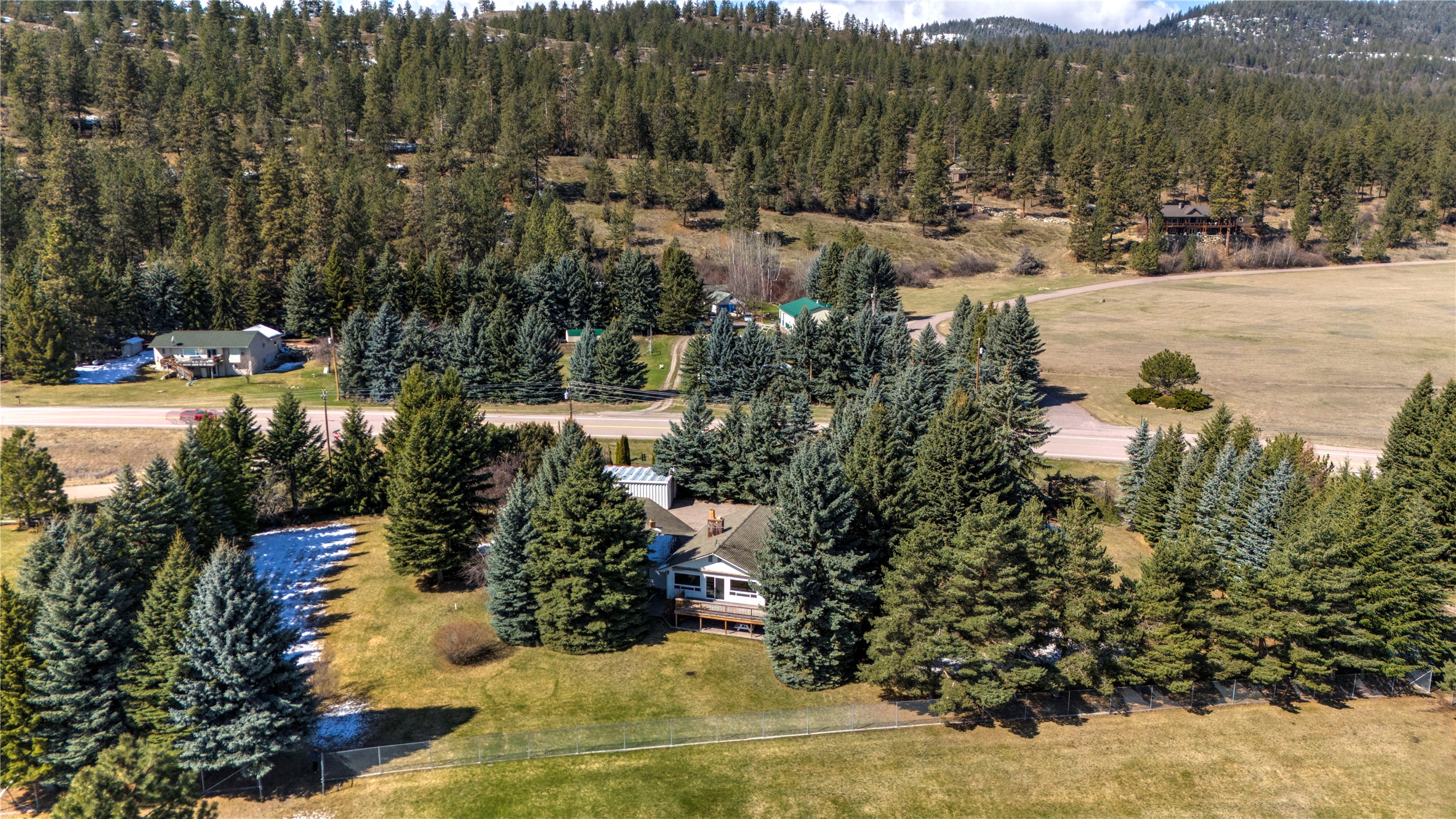Aerial view of a house nestled among evergreens, with a fenced yard and road nearby.
