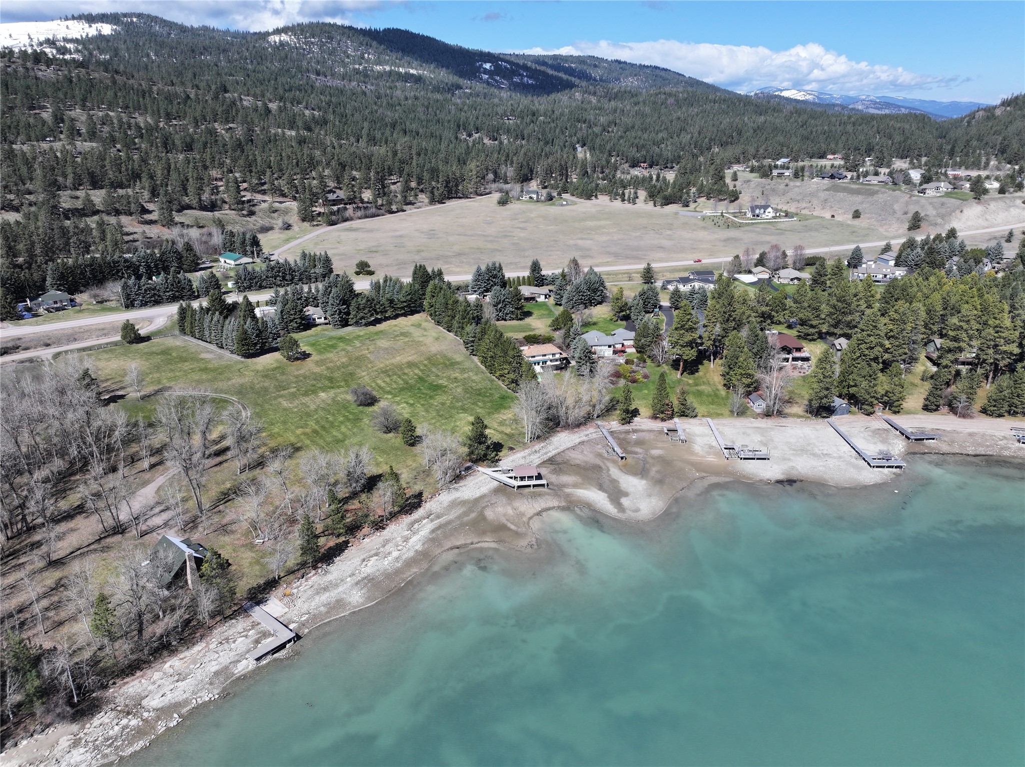 Aerial view of a wooded lakeshore with several docks, turquoise water, and forested mountains beyond.