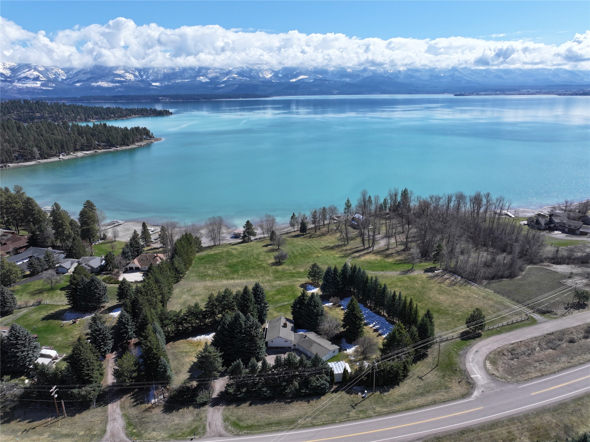 Aerial view of a lakeside home and grounds, with blue water and snow-dusted mountains beyond.