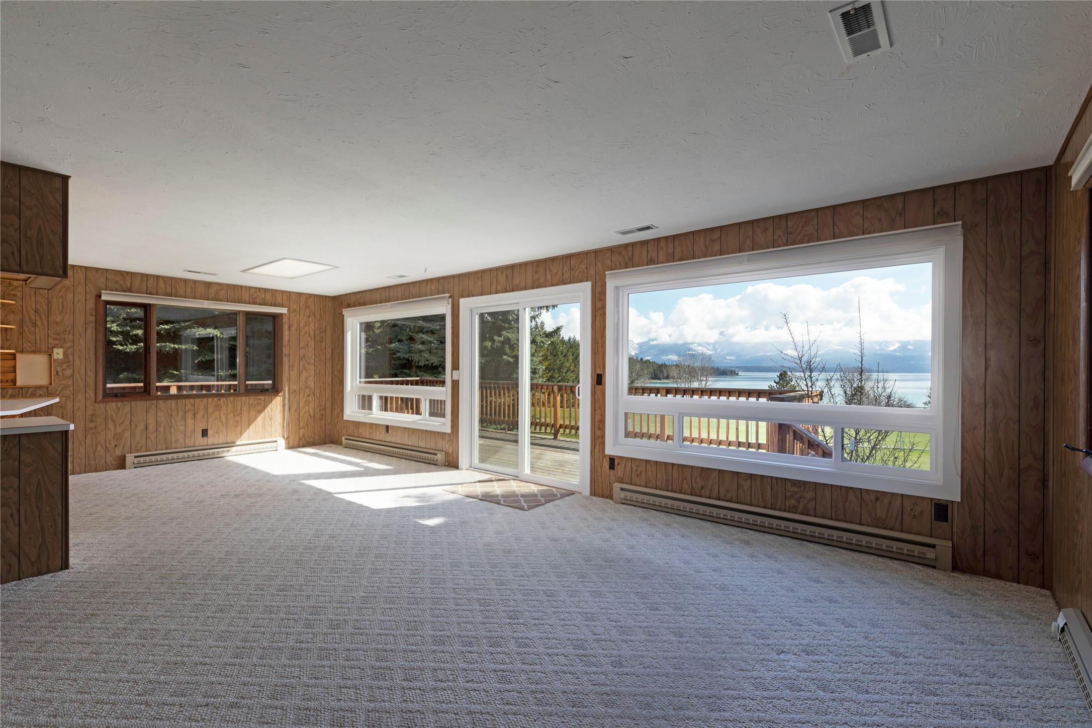 Empty wood-paneled living room with large windows overlooking a lake and distant mountains.