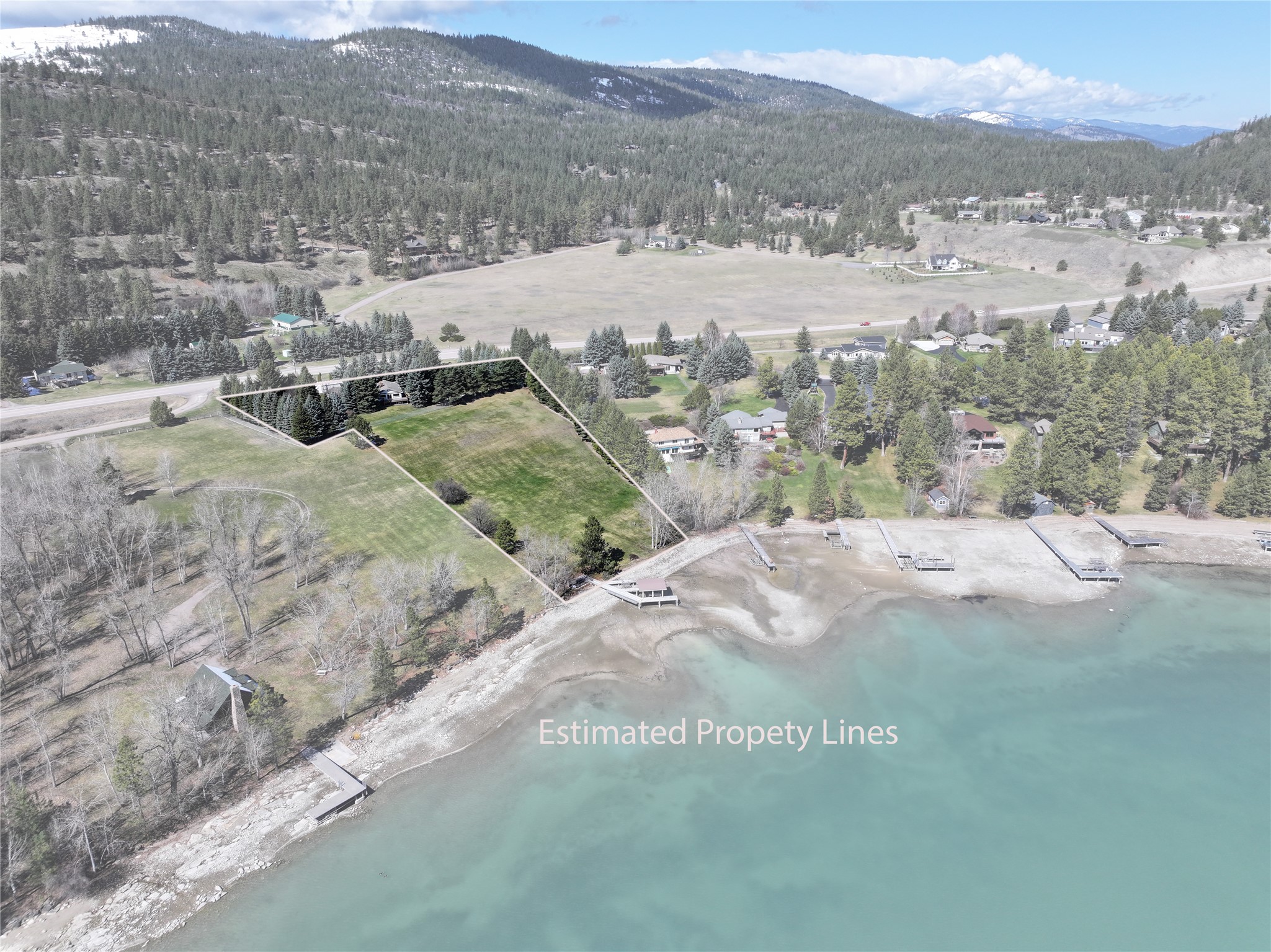 Aerial view of outlined lakefront property with several docks, turquoise water, and snow-dusted mountains beyond.