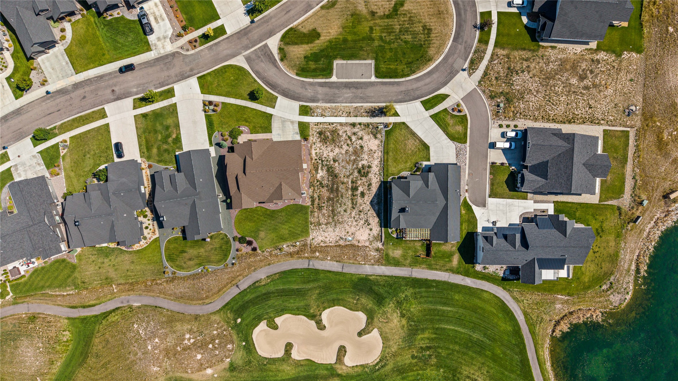 Aerial view of a vacant lot among homes, beside a golf course and water.
