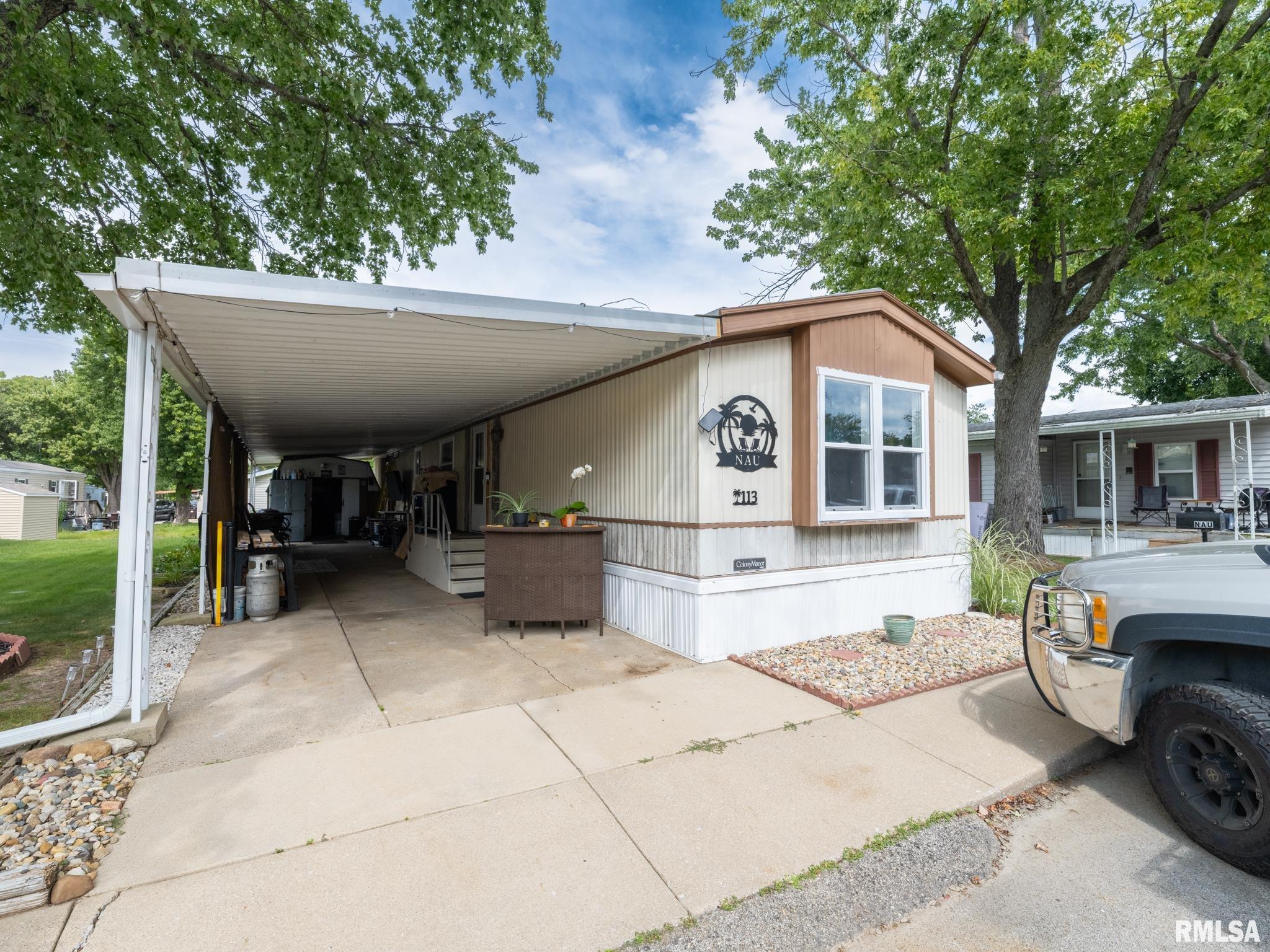 Manufactured home with a covered carport, brown trim, and a palm-tree emblem beside the front window.