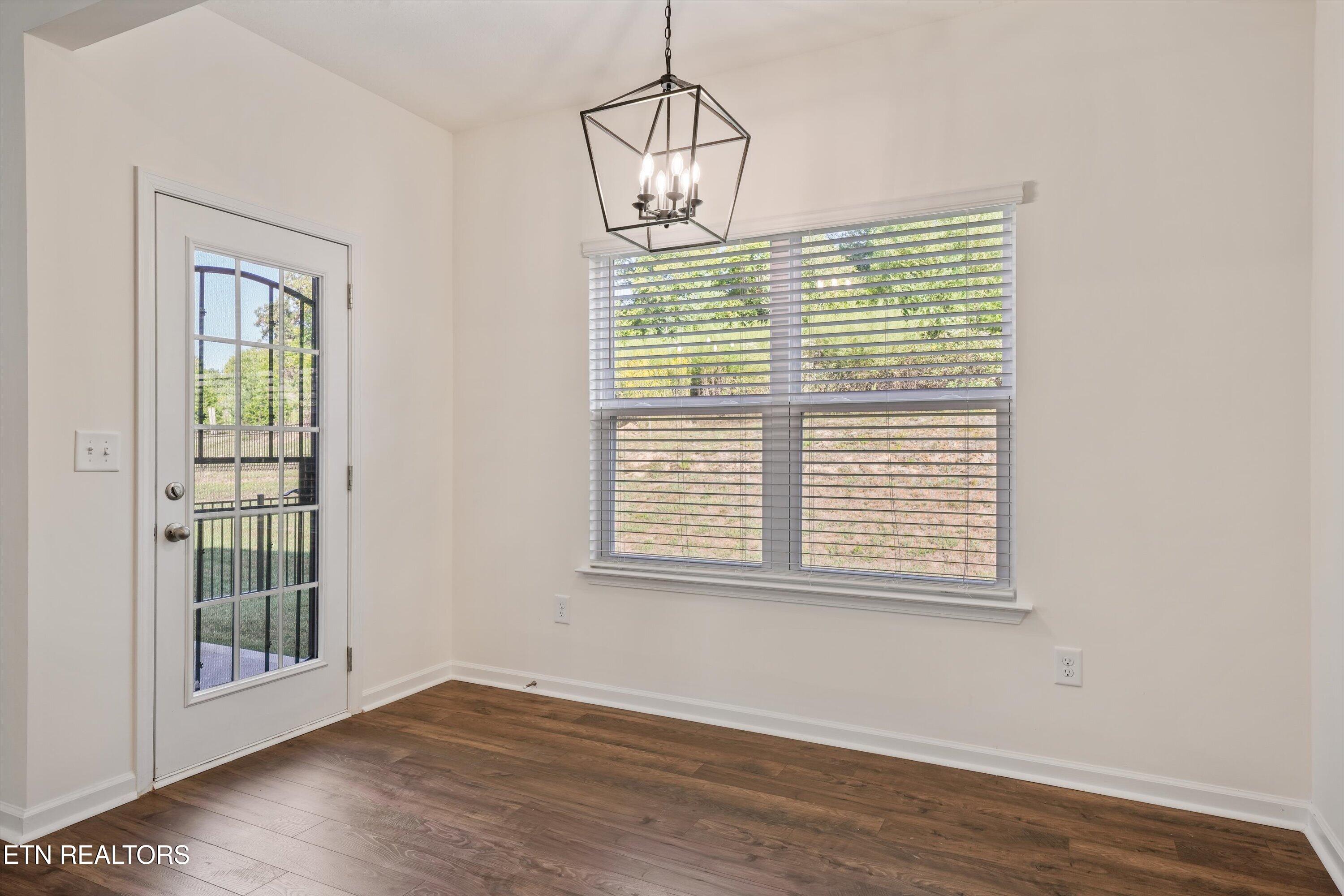 Empty room with dark wood floors, a glass exterior door, a large blinded window, and a geometric pendant light.