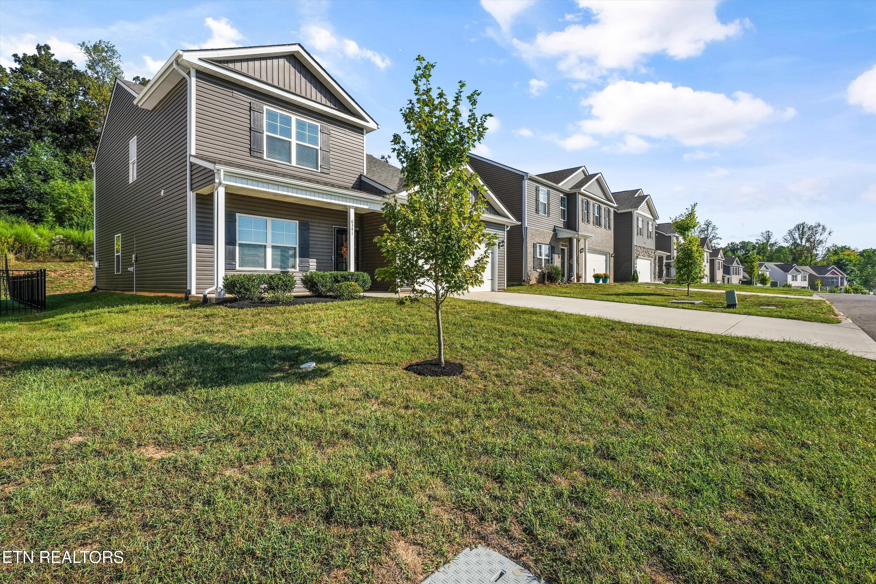 Two-story gray-sided home with a covered porch, broad lawn, and young tree in front.