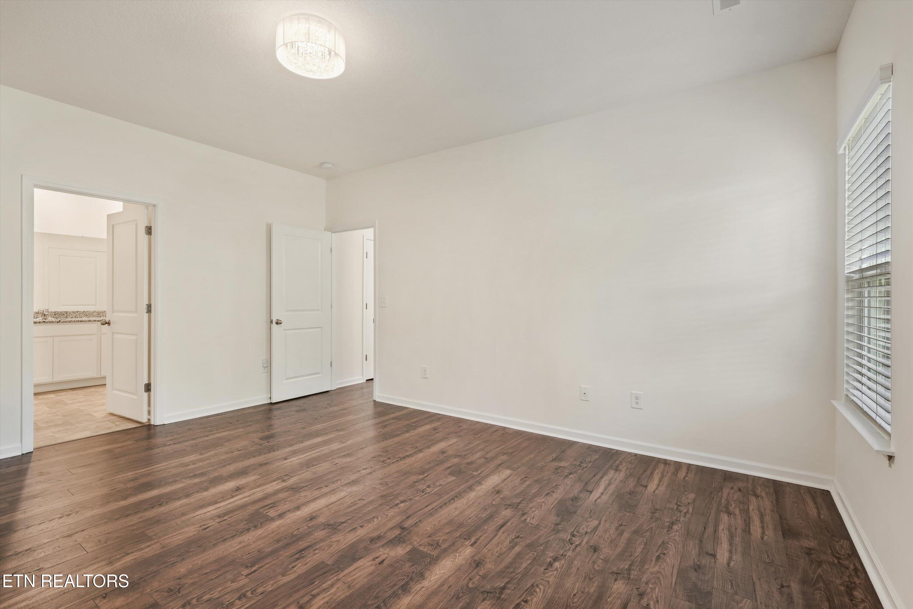 Empty room with dark wood flooring, a ceiling light, and a doorway to the kitchen.