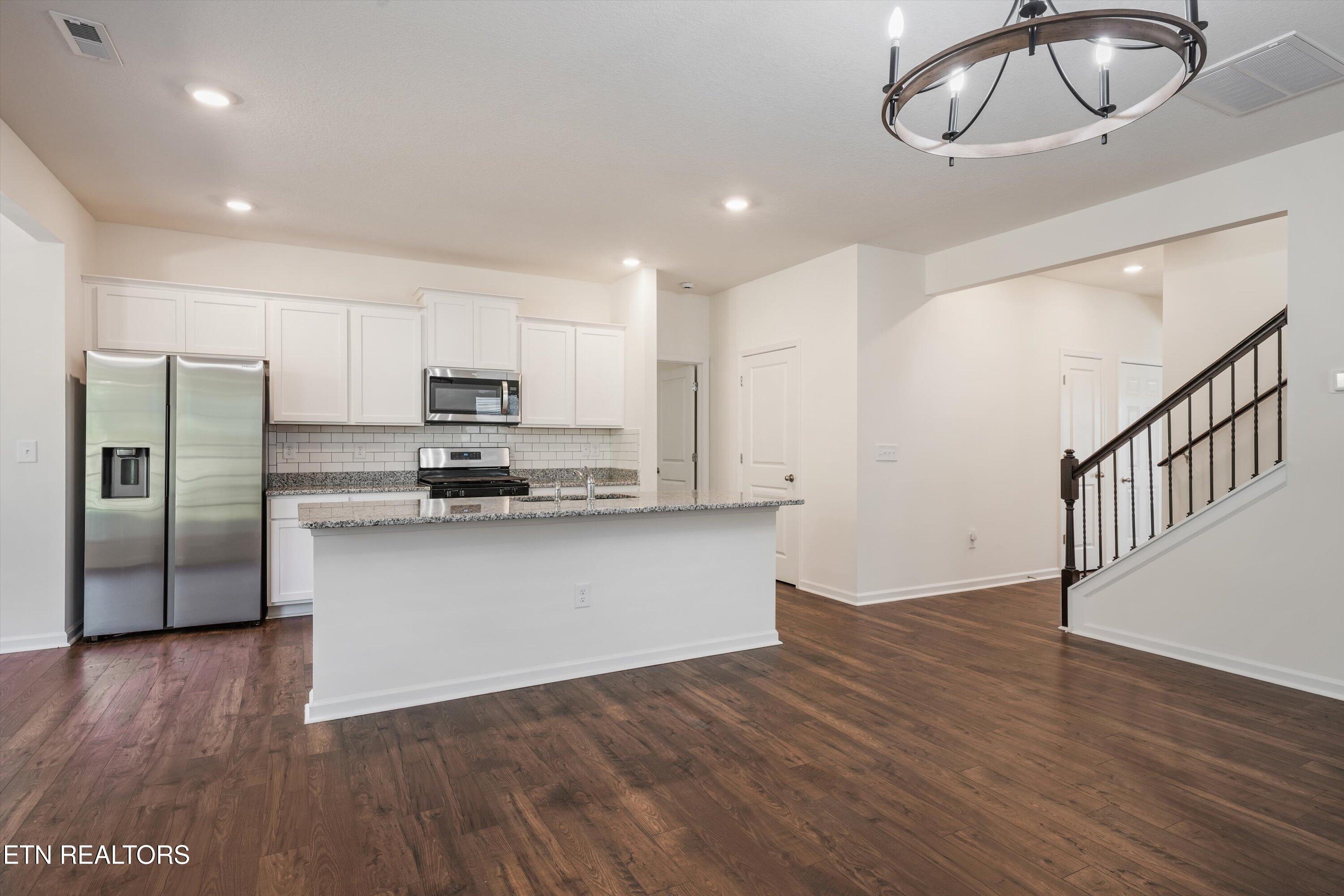 Kitchen with white cabinets, stainless steel appliances, granite island, dark wood floors, and an adjacent staircase.