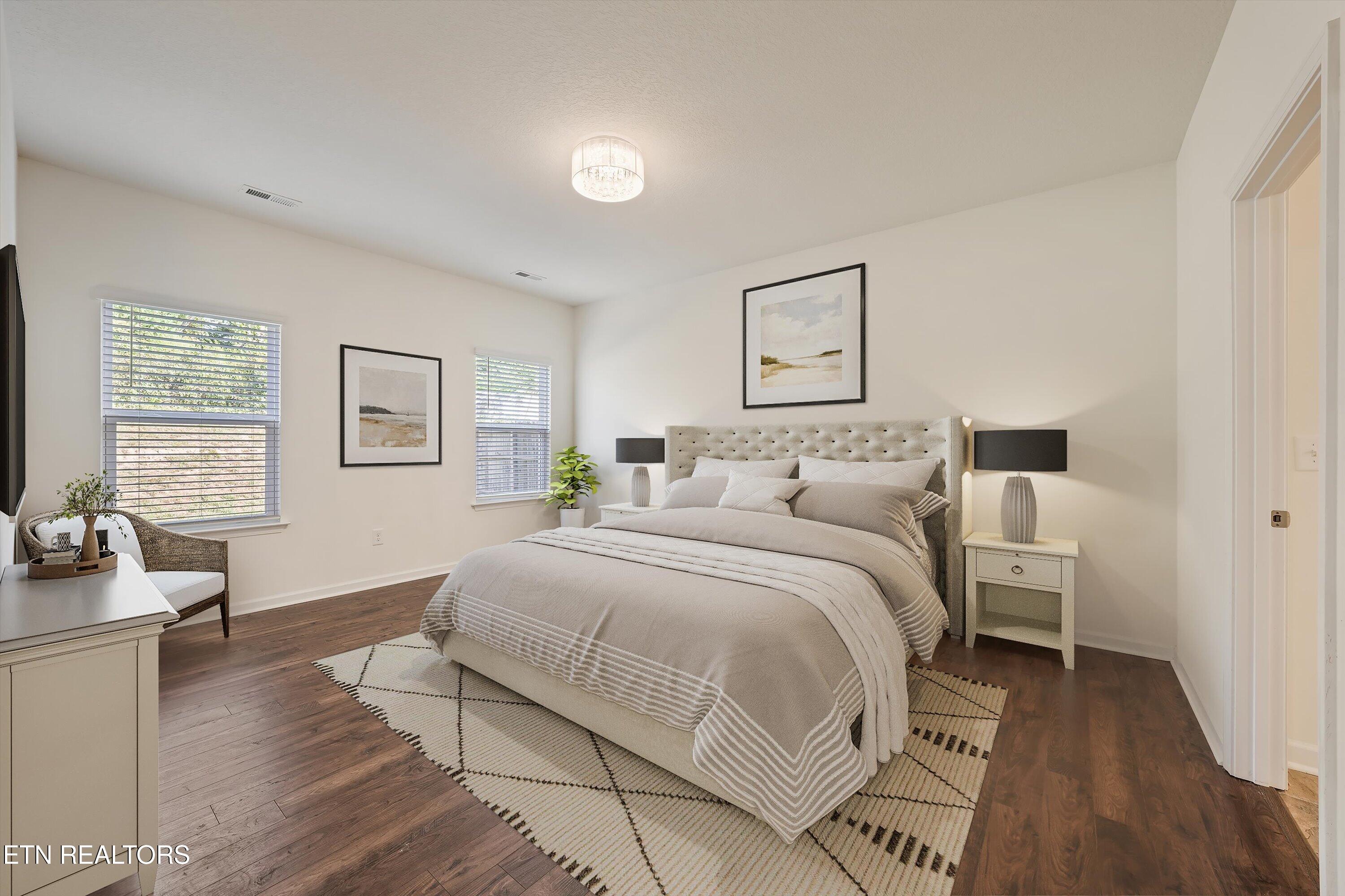 Staged bedroom with an upholstered bed, matching bedside lamps, two windows, and dark wood floors.