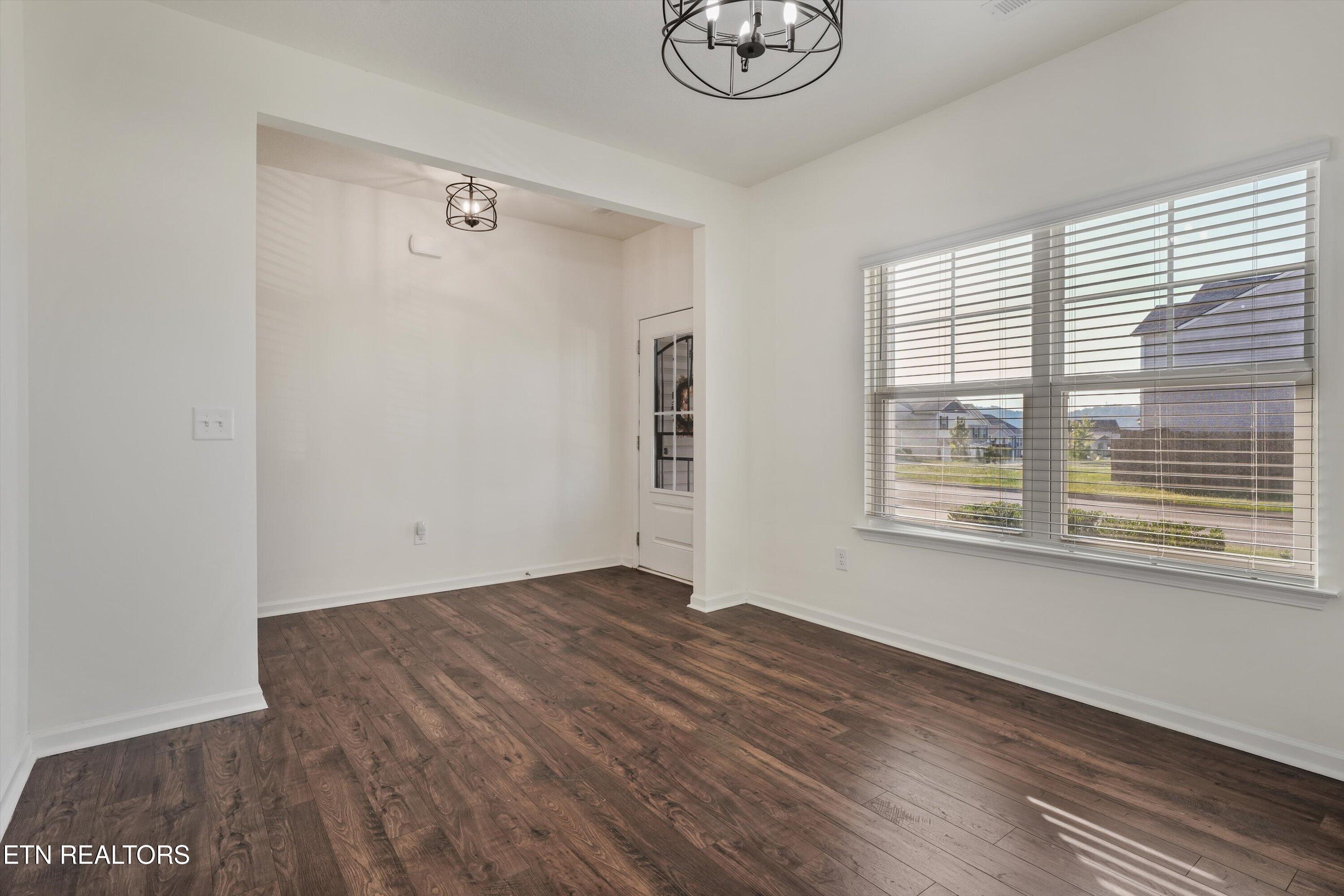 Empty room with dark wood floors, two chandeliers, and a large window overlooking neighboring homes.