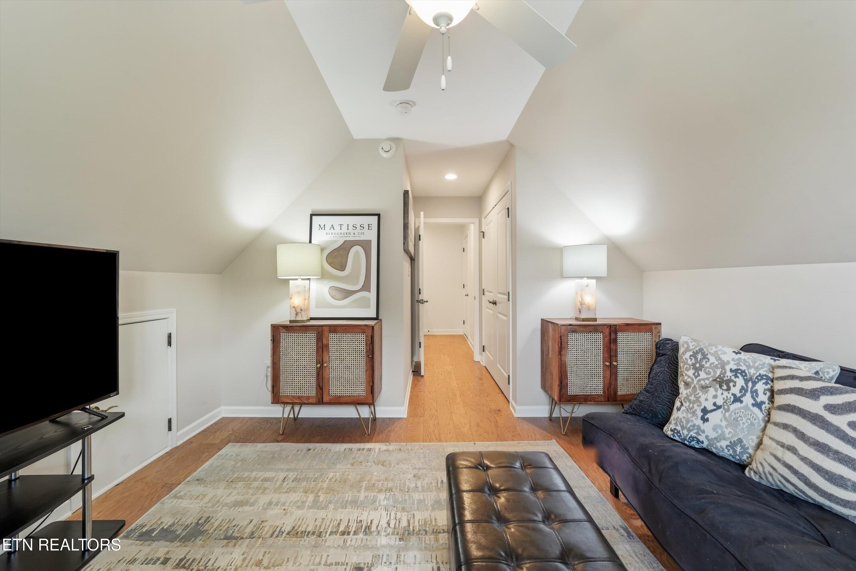 Staged living room with vaulted ceiling, hardwood floors, dark sofa, and matching wood cabinets.