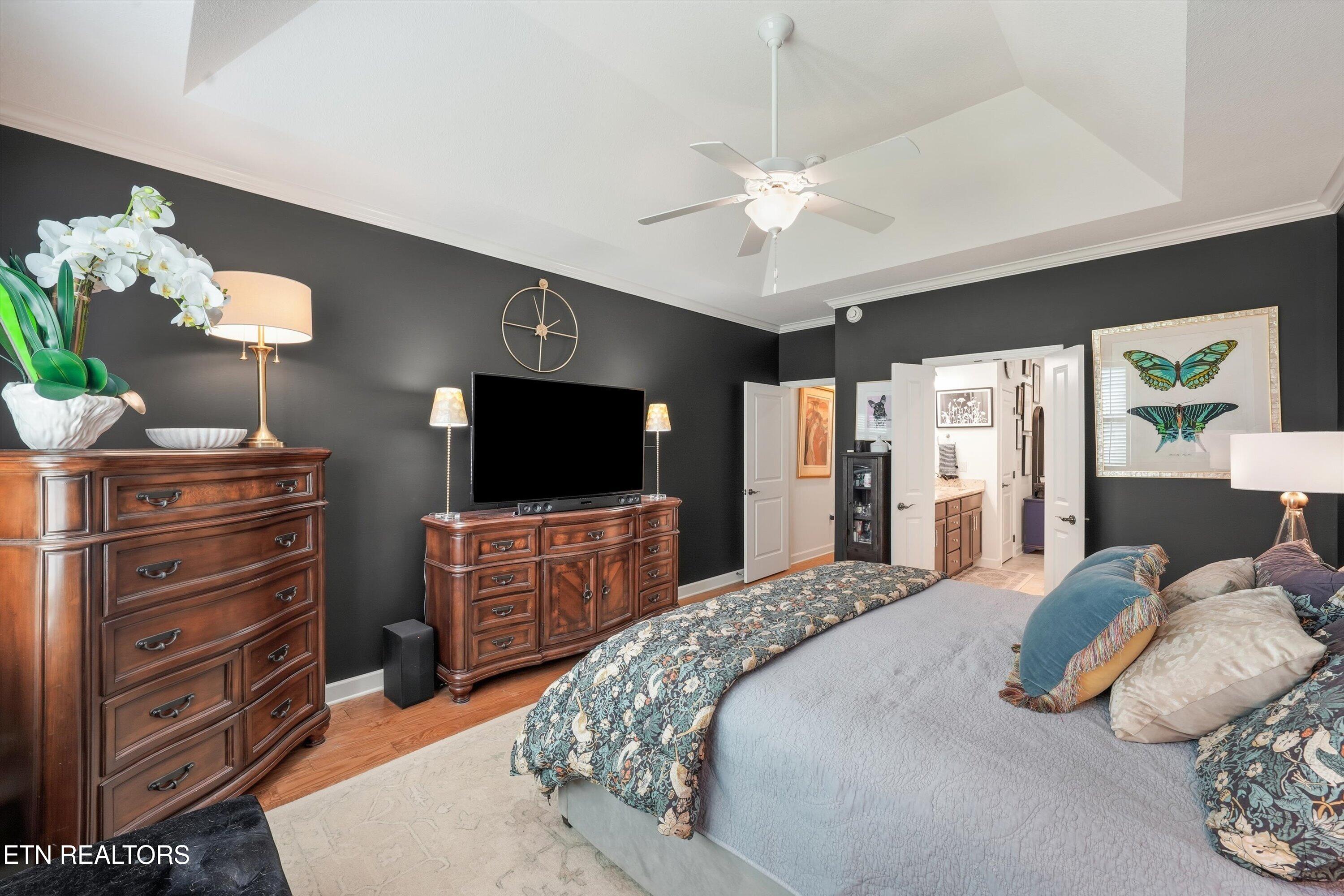 Furnished bedroom with charcoal walls, a tray ceiling, wooden dressers, and open doors to an adjoining bathroom.