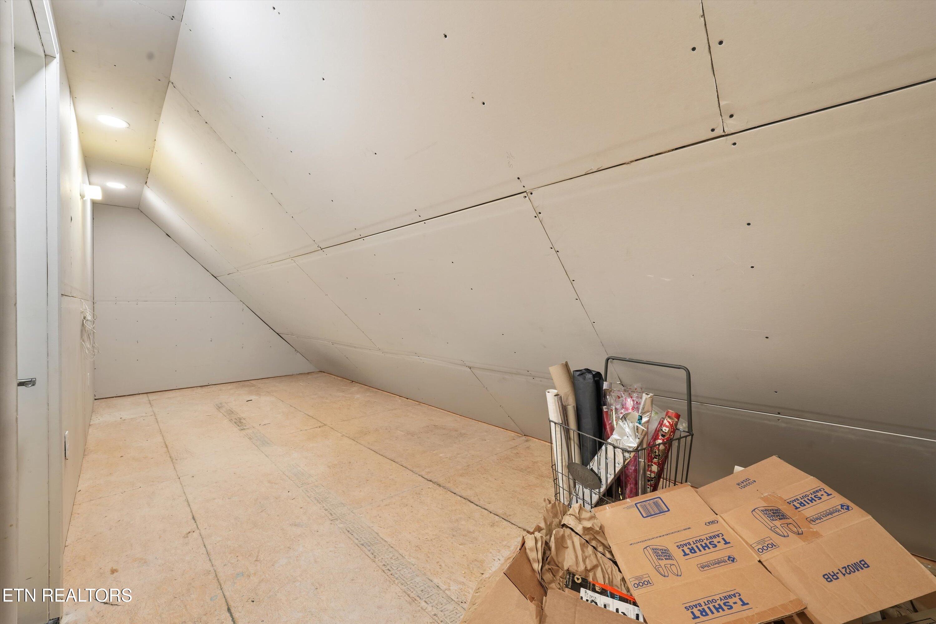 Unfinished attic with sloped drywall ceiling, plywood flooring, and boxes and supplies in one corner.