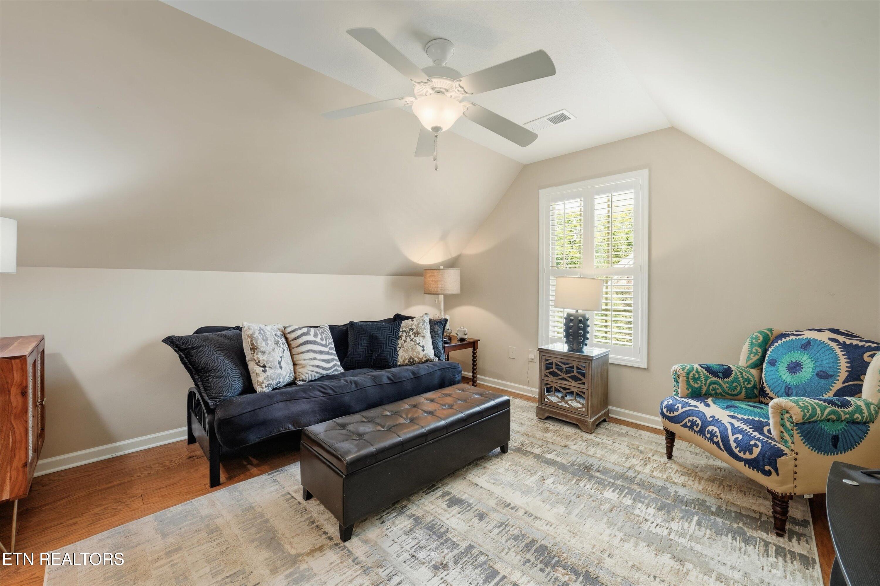 Sitting room with a dark sofa, tufted ottoman, patterned armchair, and shuttered window under a sloped ceiling.