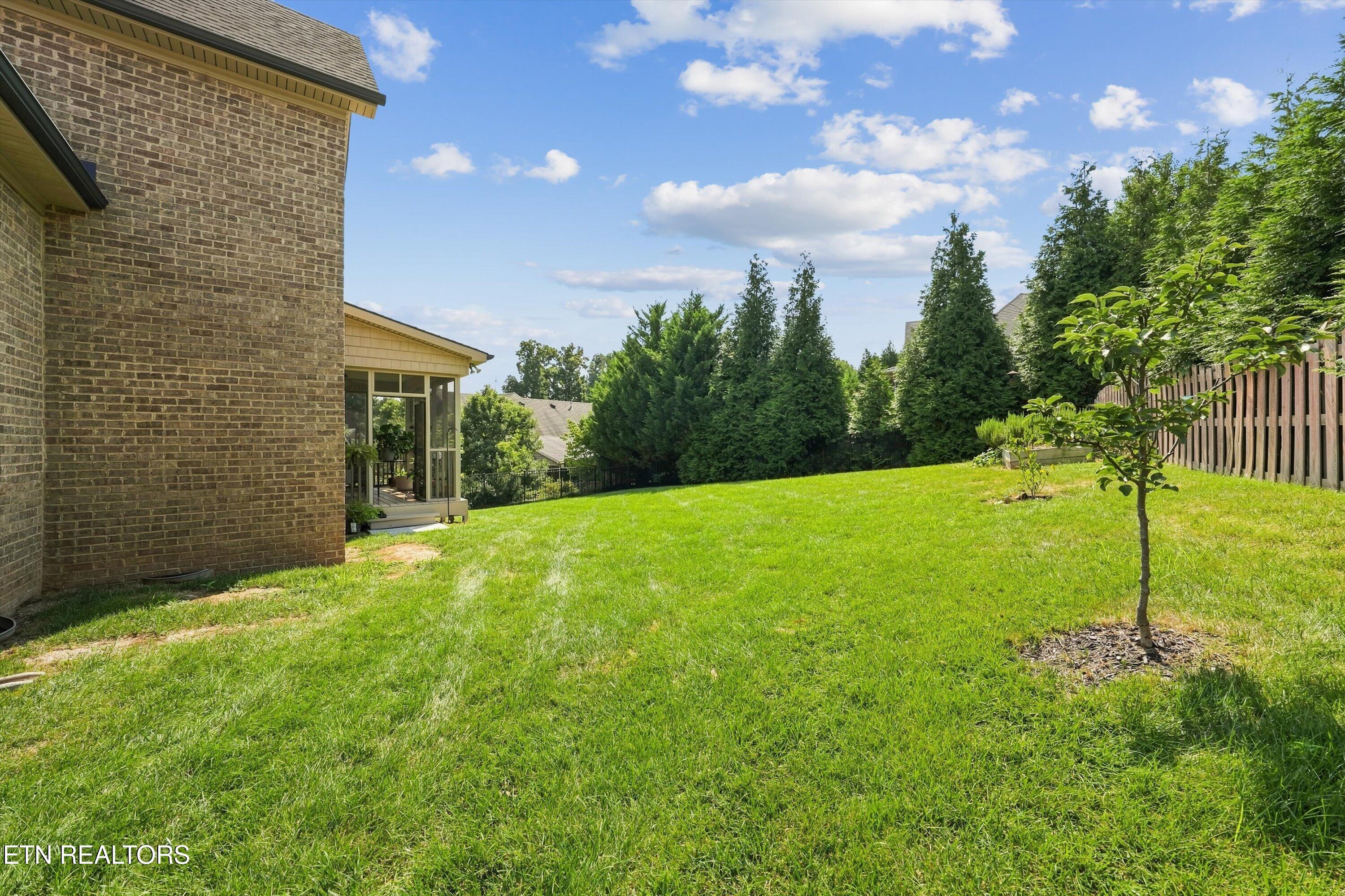 Backyard lawn with a young tree, wooden fence, and glass-enclosed porch beside a brick house.