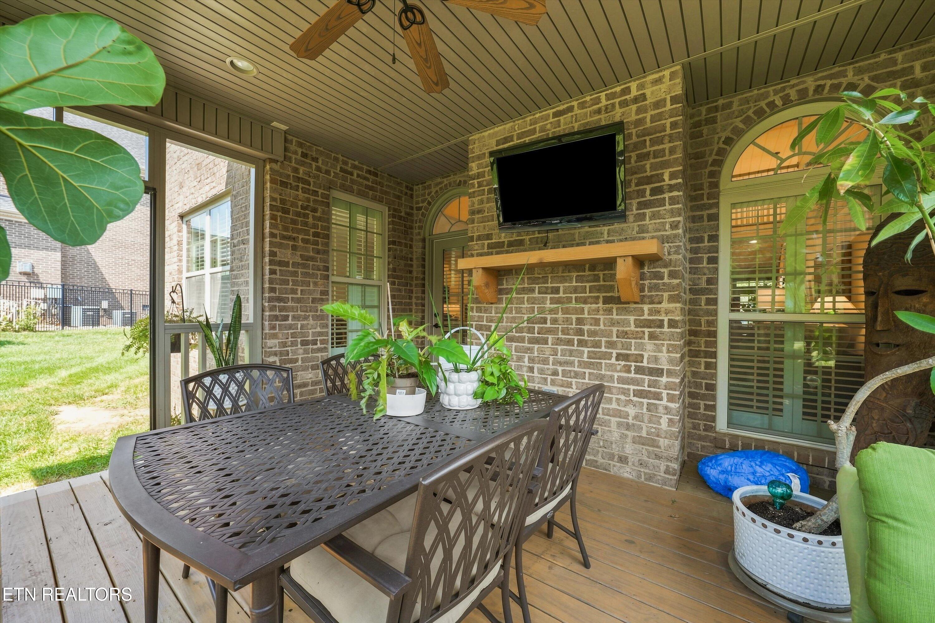 Covered brick patio with metal dining set, potted plants, and a wall-mounted television.