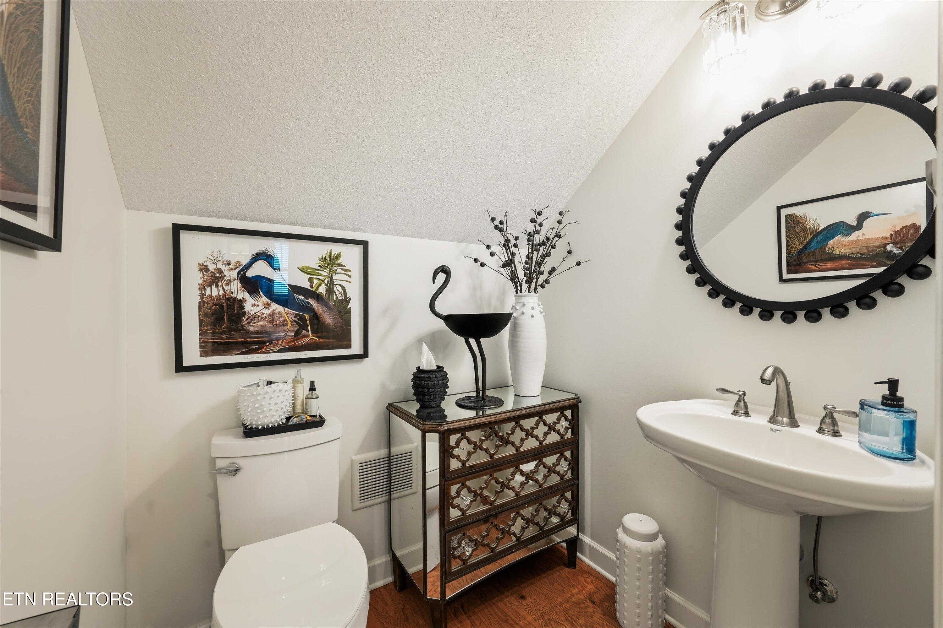 Staged bathroom with a pedestal sink, toilet, ornate mirrored chest, and large round mirror.