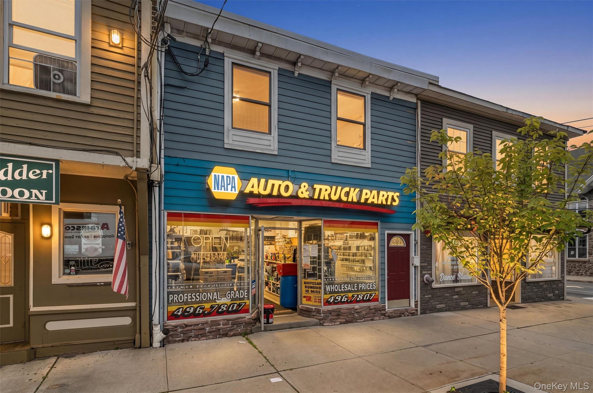 NAPA Auto & Truck Parts storefront with illuminated windows and a sidewalk tree at dusk.