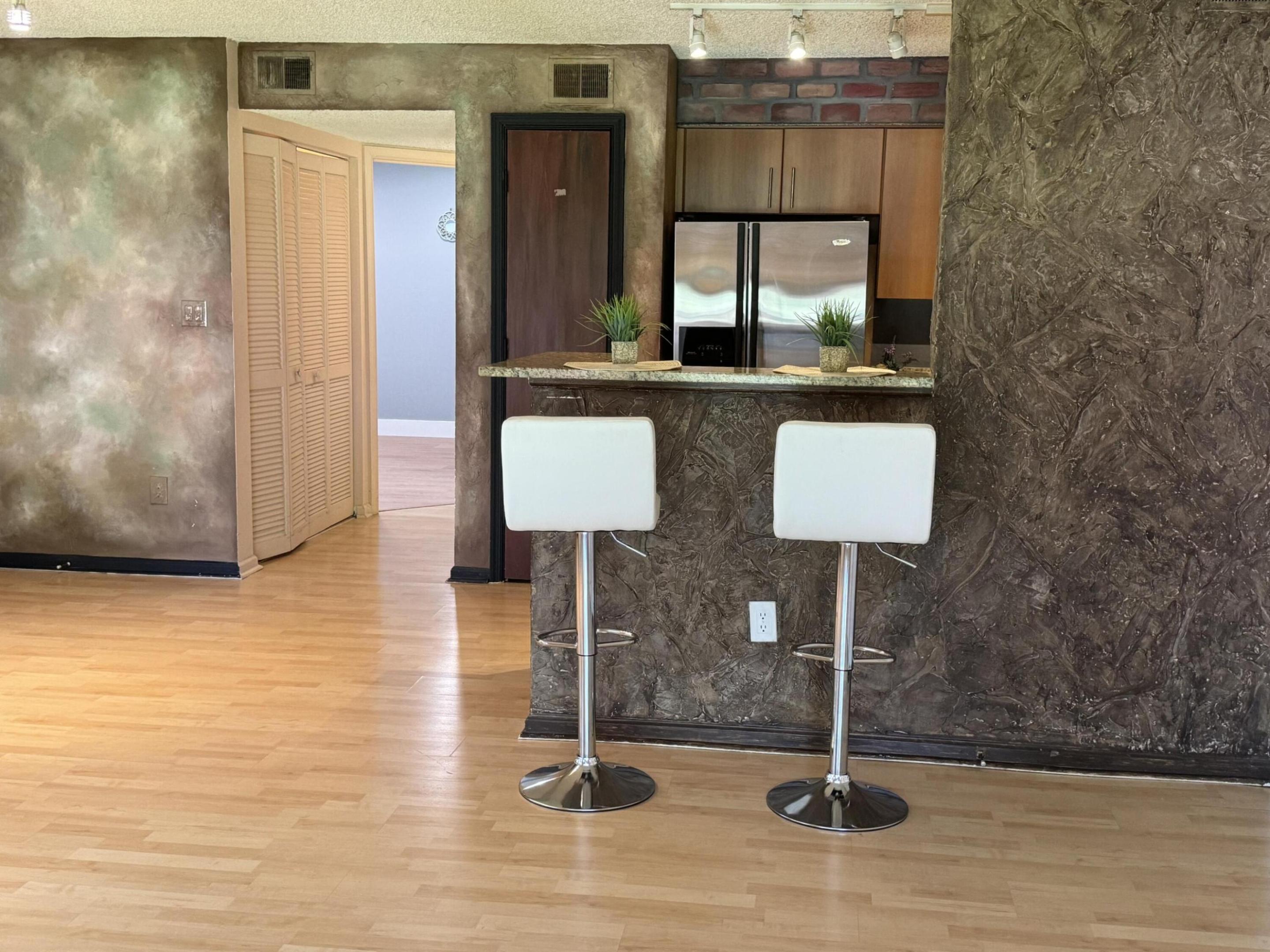 Kitchen bar with two white stools, a stainless steel refrigerator, and textured stone counter front.