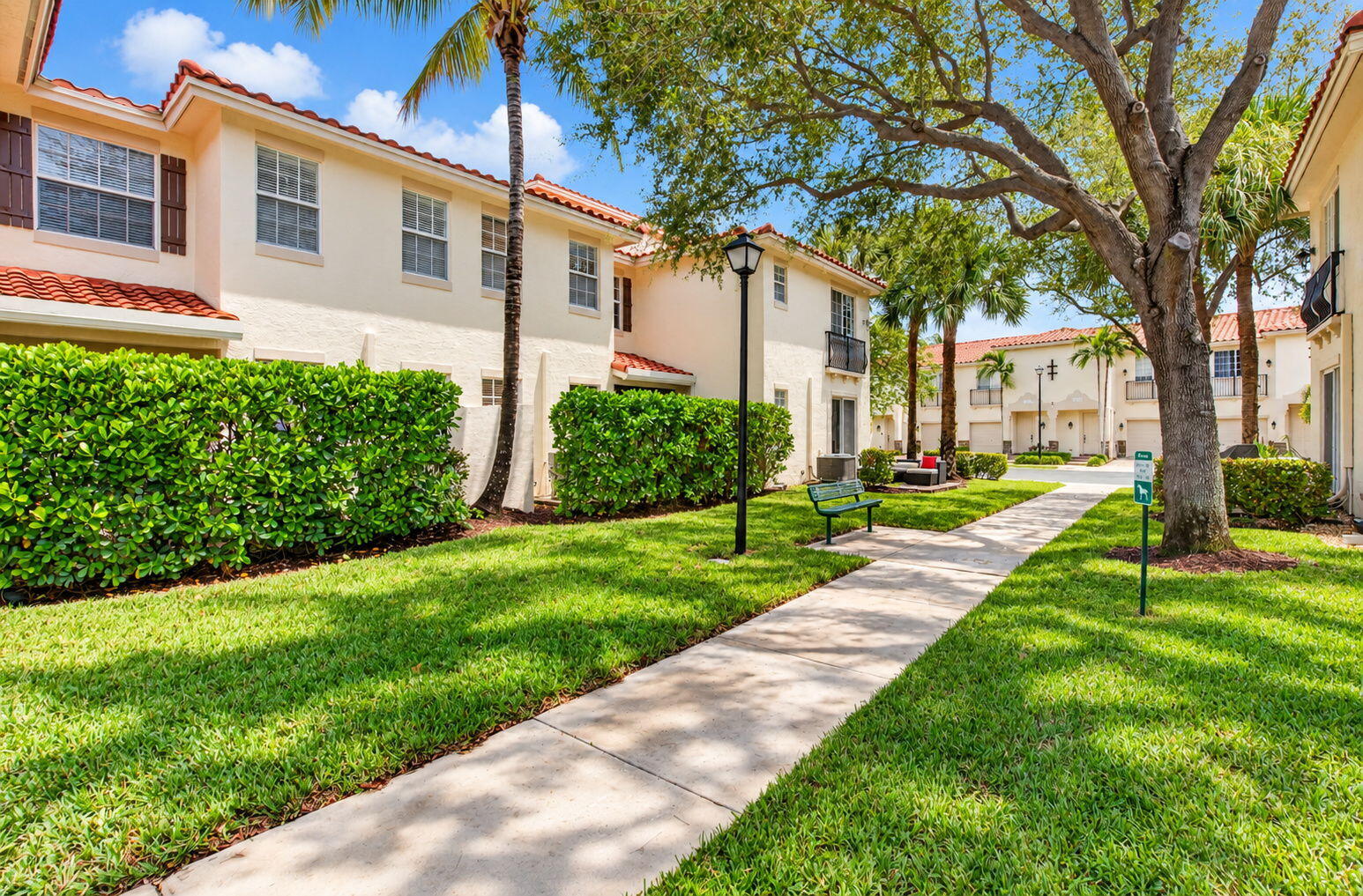Townhouse courtyard with a paved walkway, trimmed hedges, palms, and a large shade tree.