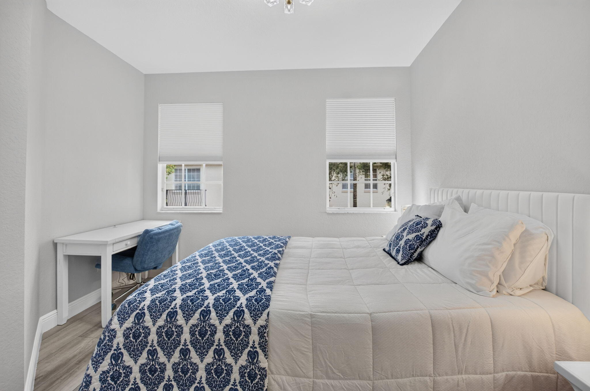 Furnished bedroom with a white bed, navy patterned throw, and desk beneath two windows.