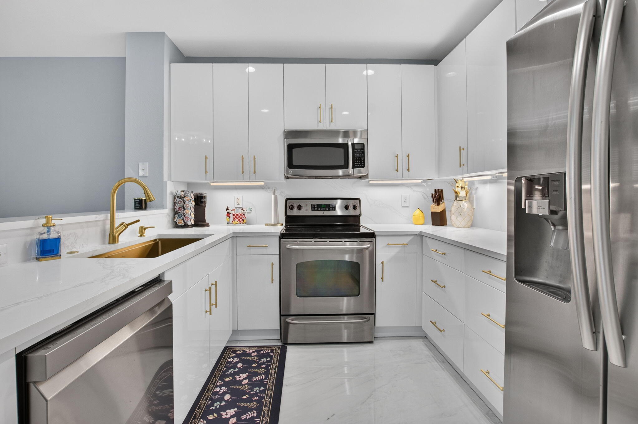 White kitchen with stainless steel appliances, marble-look counters, gold fixtures, and a floral runner.