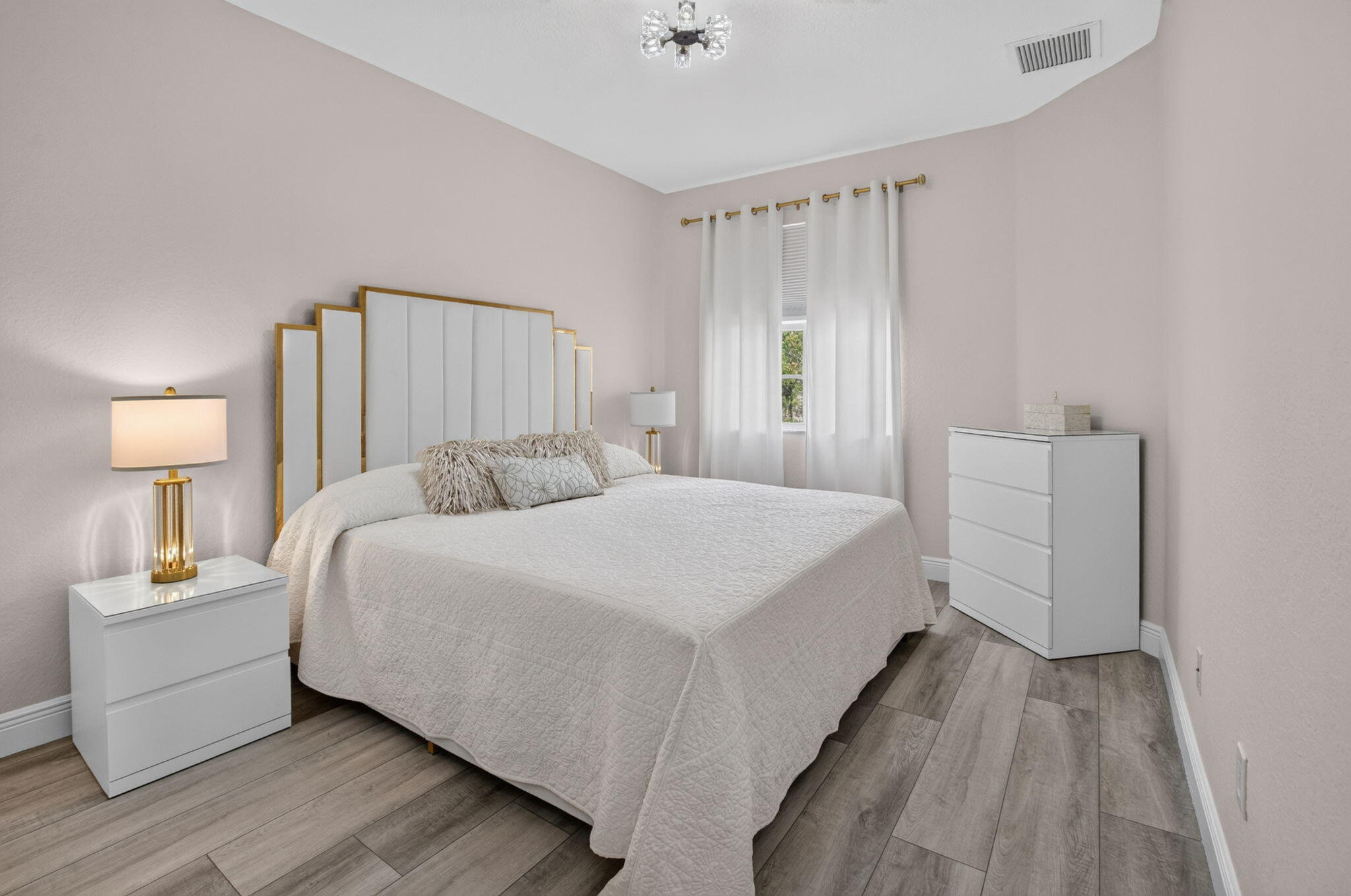 Bedroom with a white quilted bed, gold-trimmed headboard, and gray wood-look flooring.