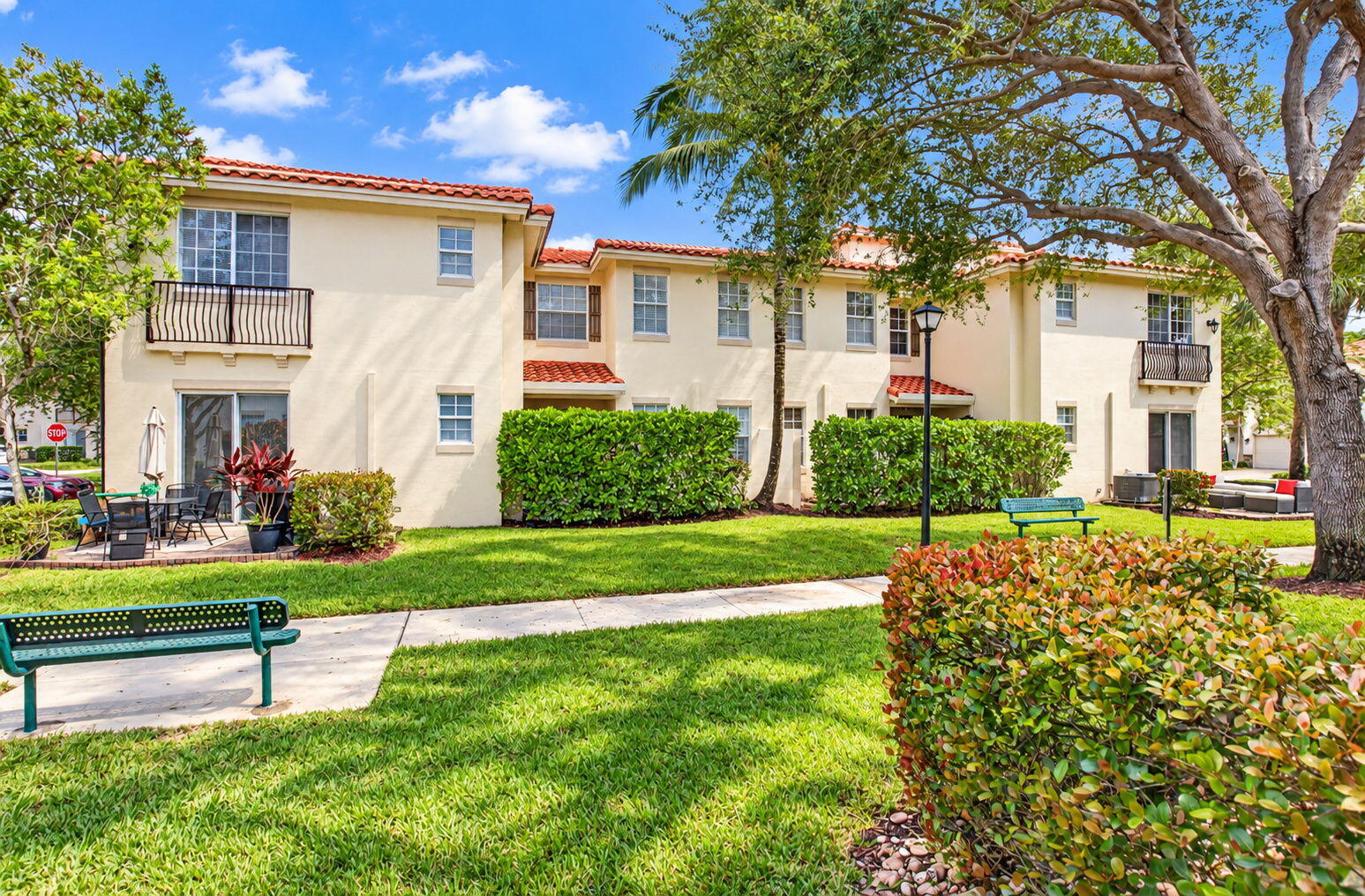 Two-story cream condominium building with red tile roofs and balconies behind a landscaped lawn and benches.